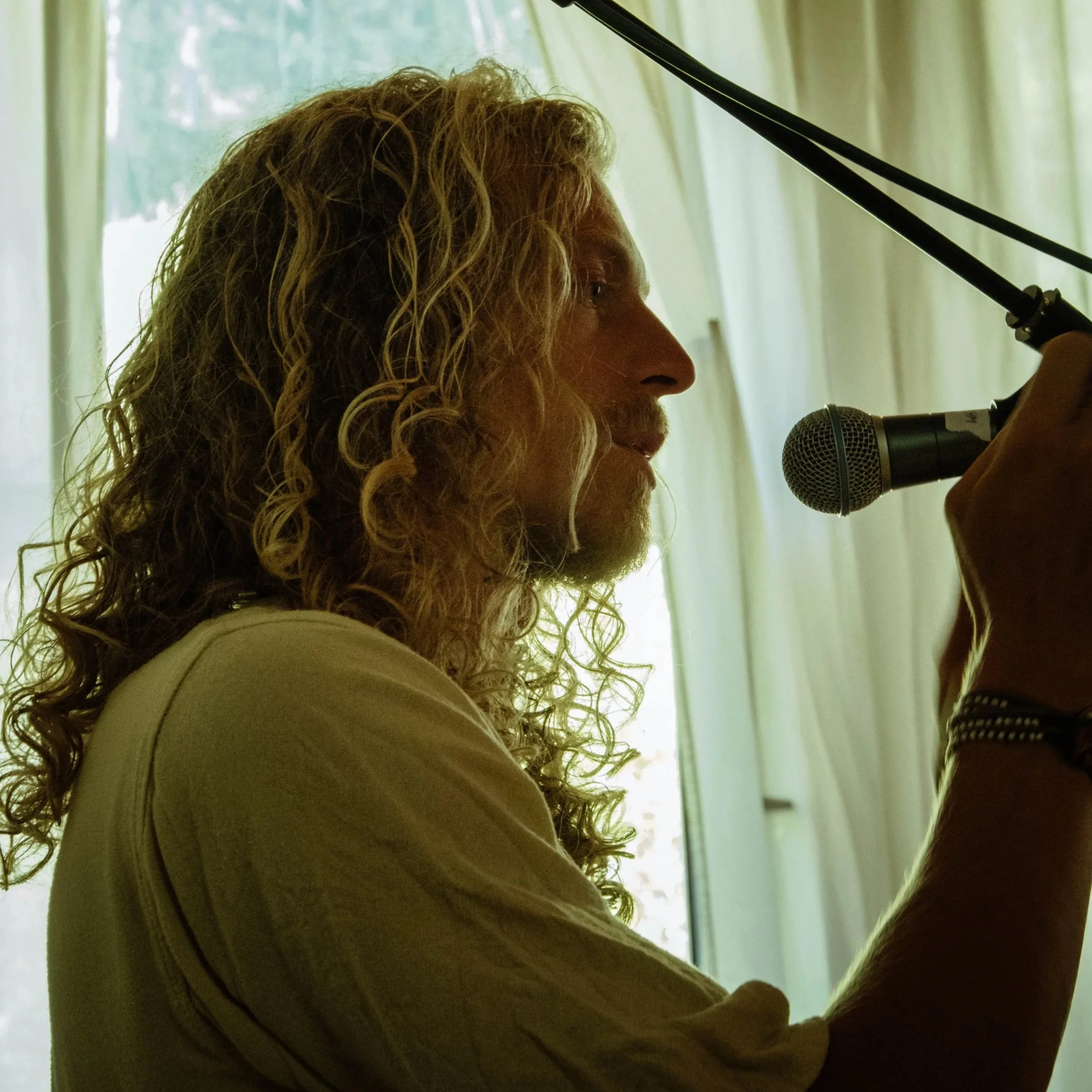 A man with long curly blond hair and a beard speaking into a microphone in front of a light-colored curtain.