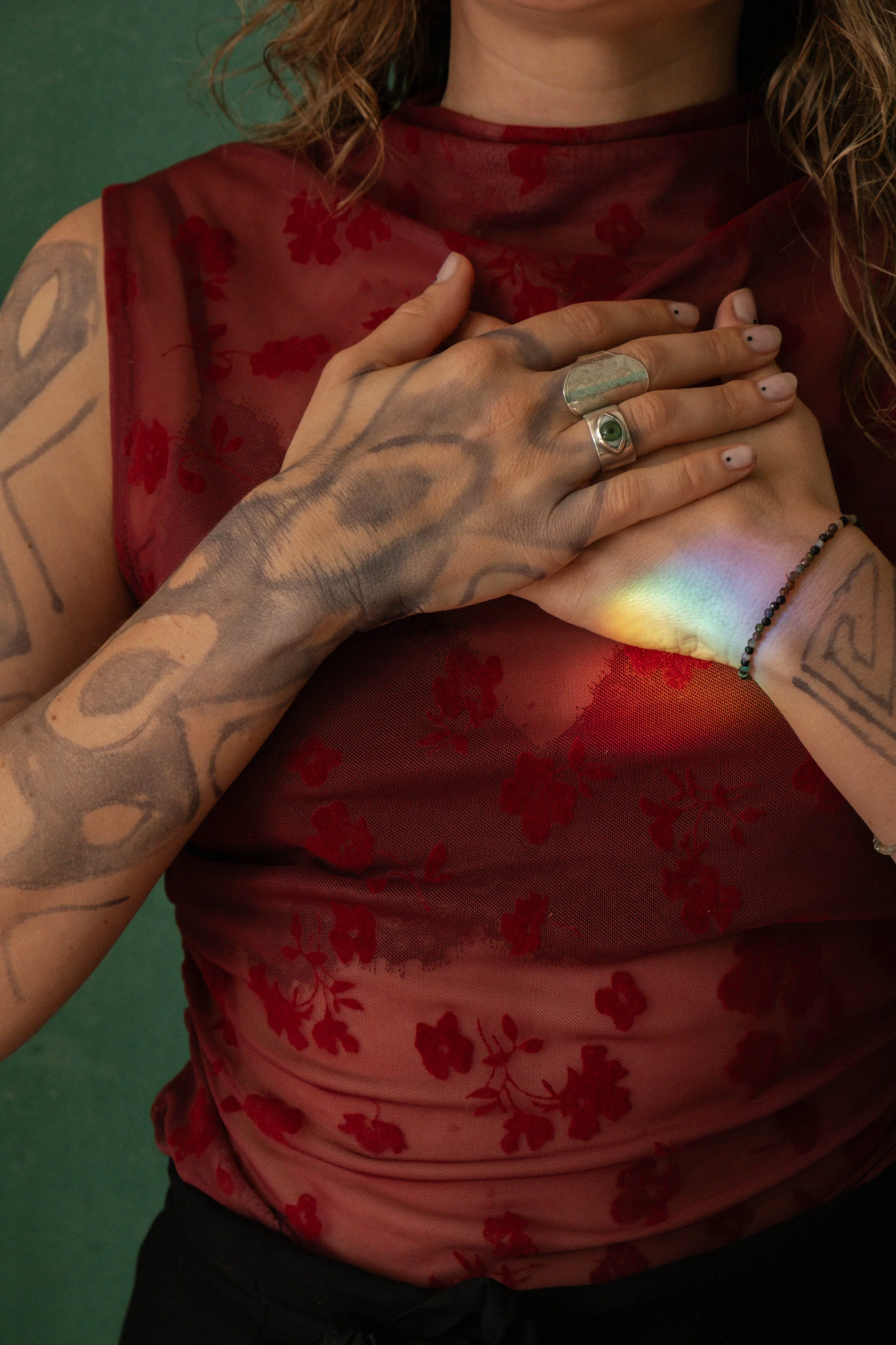 Close-up of a woman with tattoos on both arms and hand, wearing rings and a beaded bracelet, with a rainbow reflection on her wrist, touching her chest with her left hand, wearing a sheer red top with floral pattern.