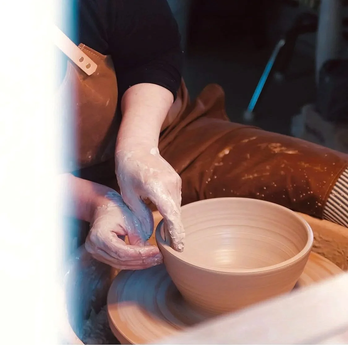 Person shaping a cup on a pottery wheel, wearing a brown apron and black shirt, hands covered in clay.
