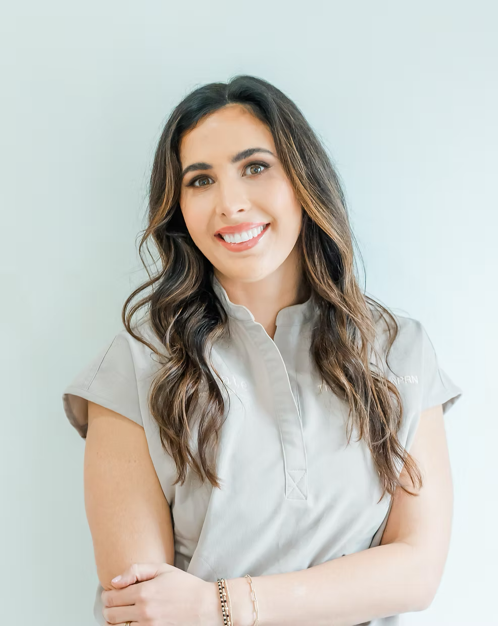 A woman with long wavy brown hair smiling at the camera, wearing a light gray collared shirt with short sleeves, standing against a plain light background.