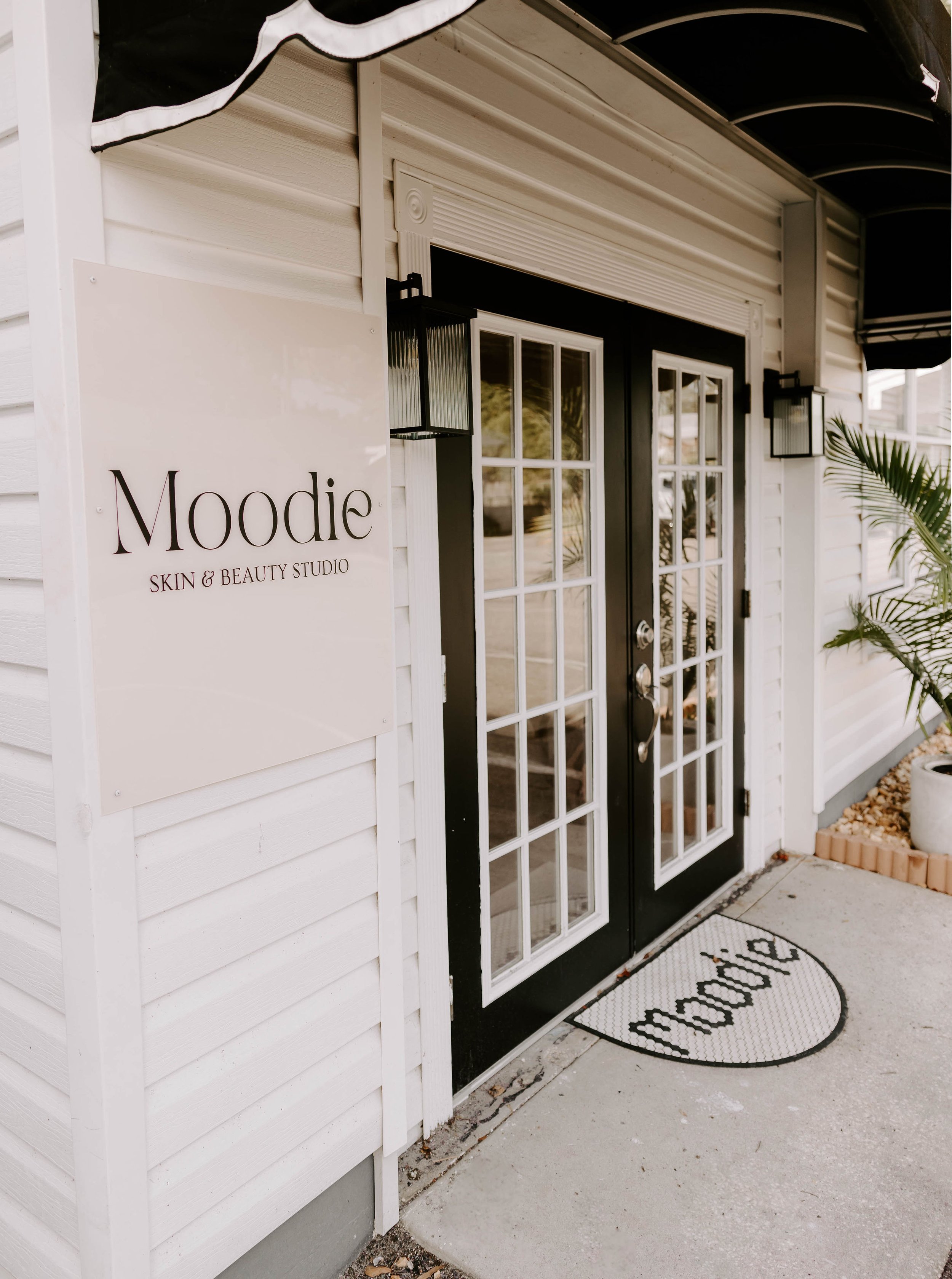 The entrance of Moodie Skin & Beauty Studio featuring double glass doors with black frames and a white sign with black text on the wall to the left. There is a semi-circular doormat with the word 'moodie' written on it in front of the door, and a pot