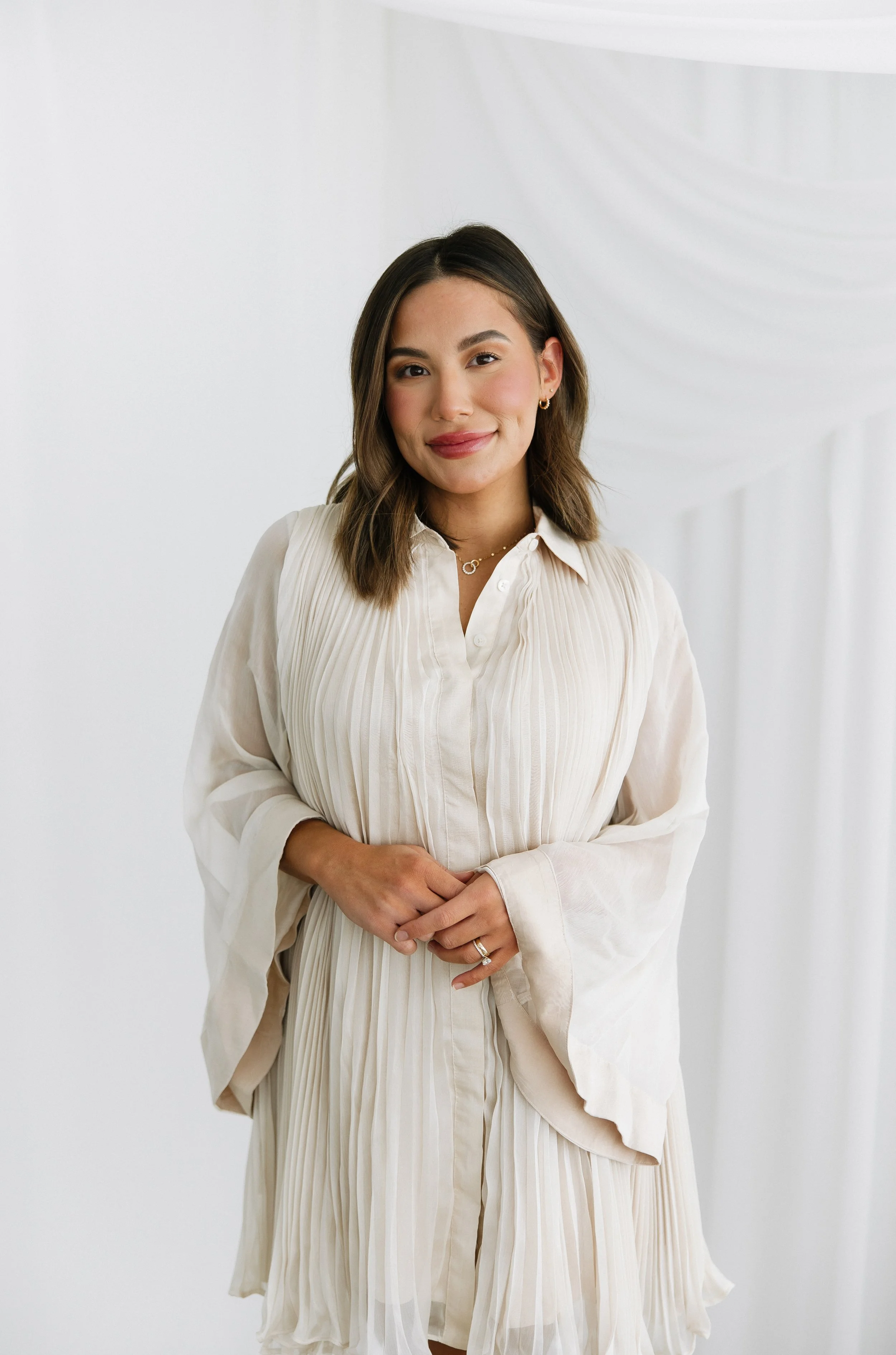 A young woman with shoulder-length brown hair, wearing a cream-colored pleated dress, standing in front of a white backdrop with sheer white curtains, smiling softly at the camera.