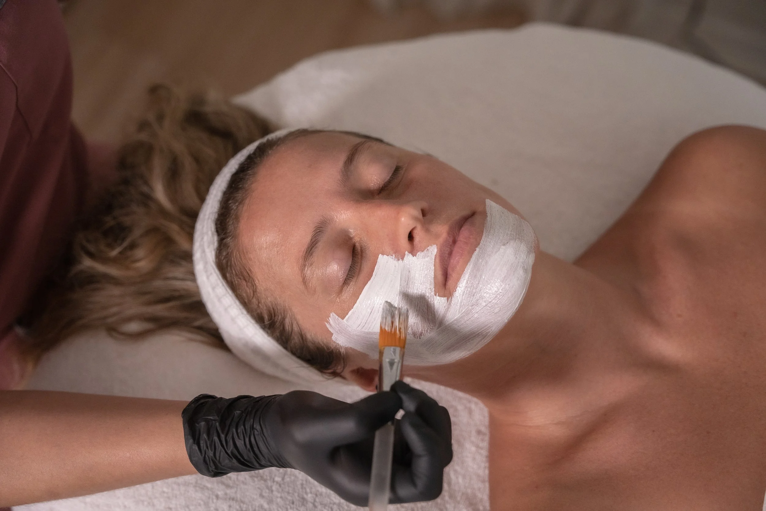 Woman receiving a facial treatment with white face mask in a spa or beauty clinic, lying on a bed with eyes closed, wearing a white headband.