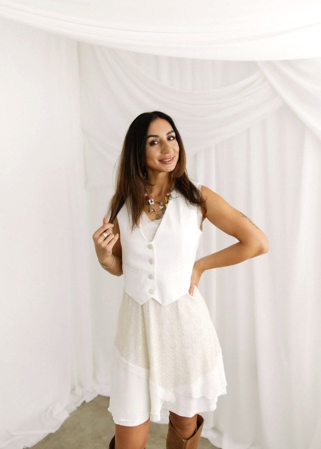 Woman wearing a white vest and skirt, standing in front of a white backdrop with draped fabric, smiling and holding a strand of hair.
