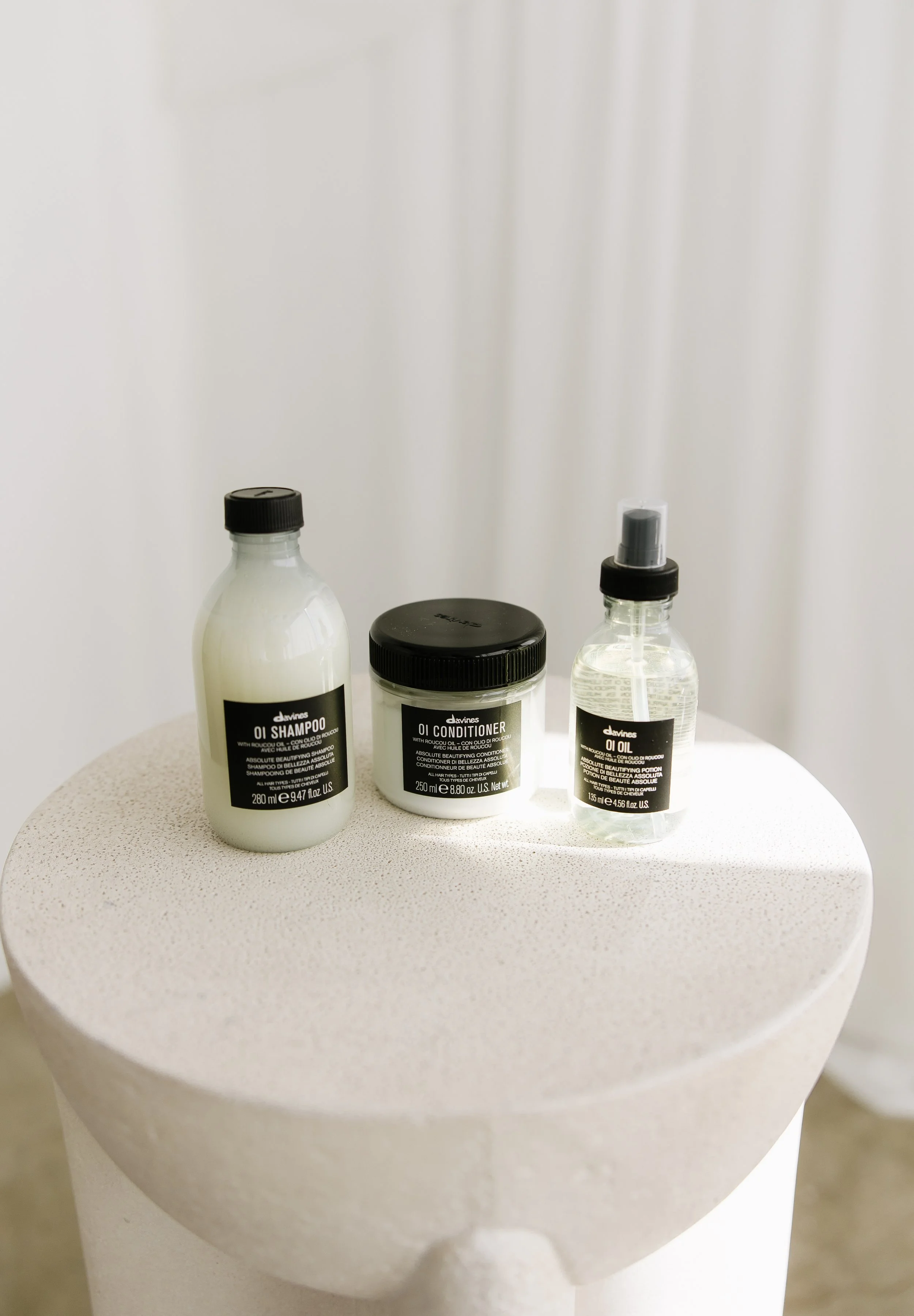 Three bottles of beauty products on a white textured round surface, including shampoo, conditioner, and oil.