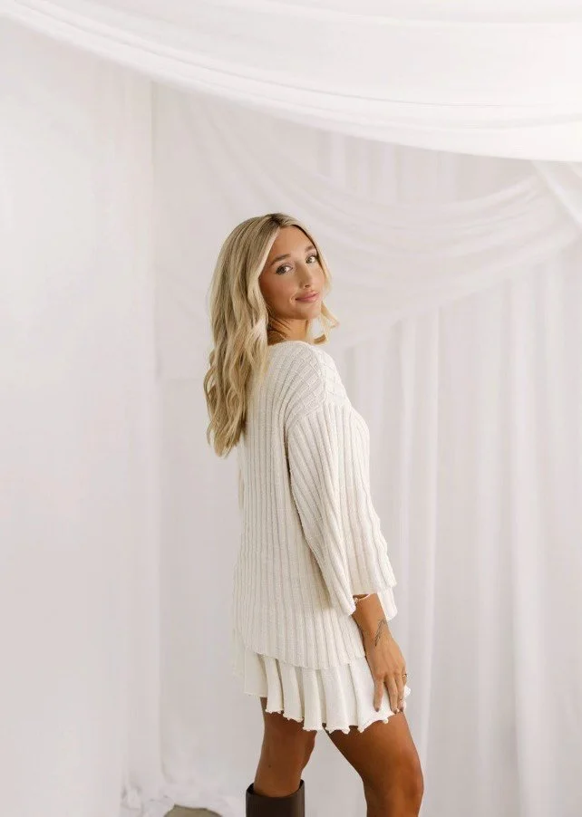 A woman with blonde hair in a white sweater and skirt, standing in front of a white draped backdrop, looking over her shoulder.