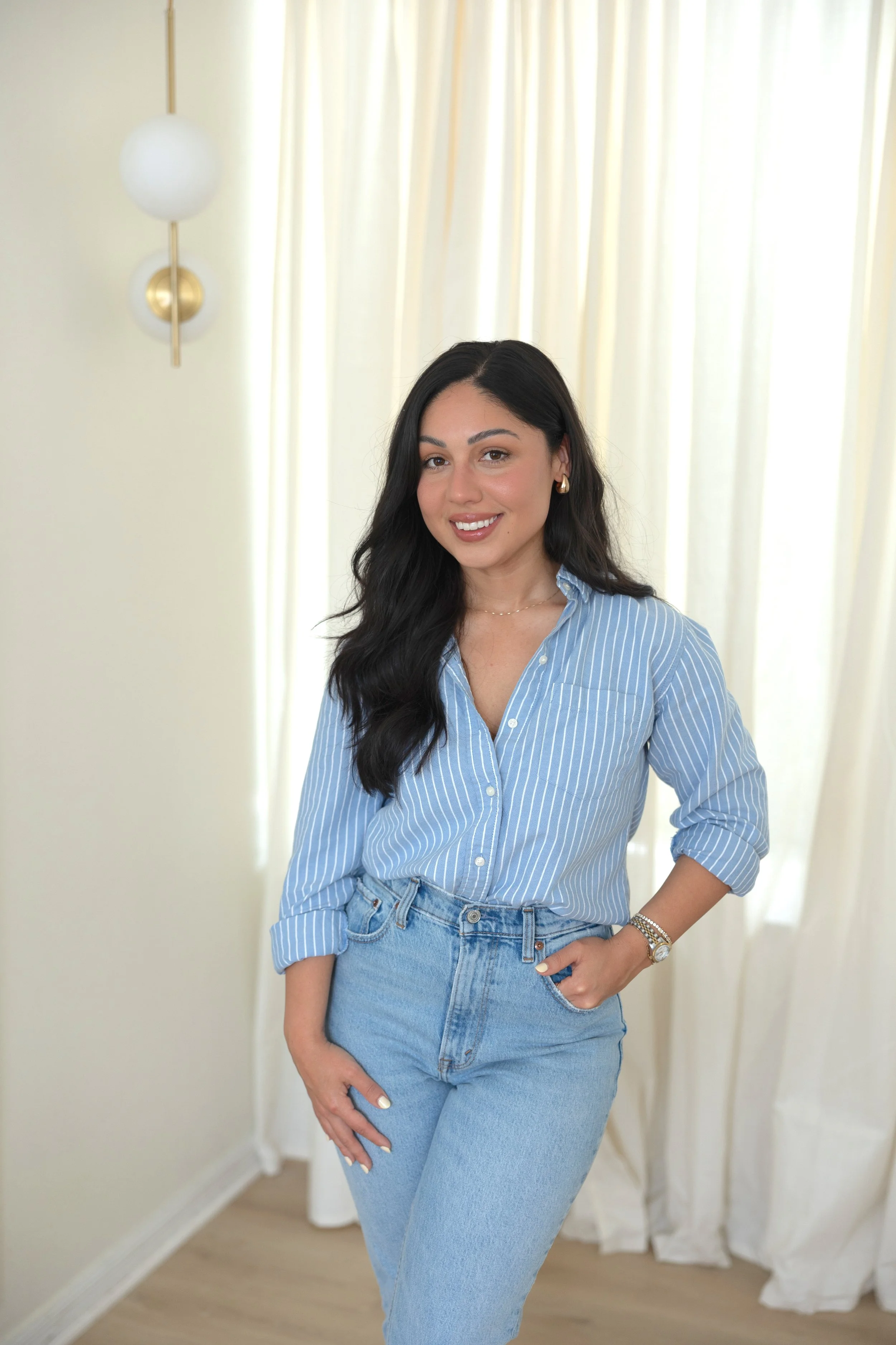 A young woman with long dark hair, wearing a blue and white striped button-up shirt and light blue jeans, standing indoors near white curtains and a modern wall sconce, smiling at the camera.