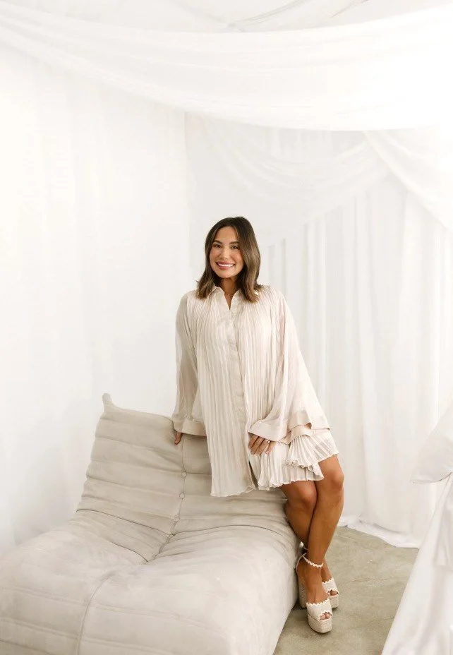 A woman in a light-colored dress sitting on a white sofa in a room with white drapery.