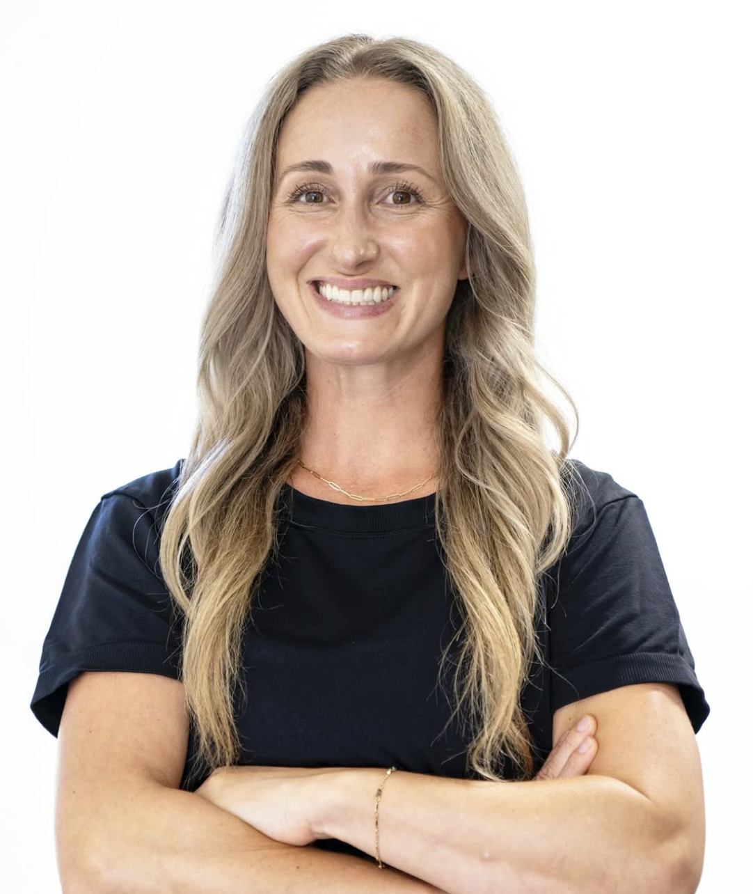 A woman with long, wavy blonde hair, smiling, wearing a black t-shirt, with arms crossed and a simple gold necklace and bracelet, against a plain white background.