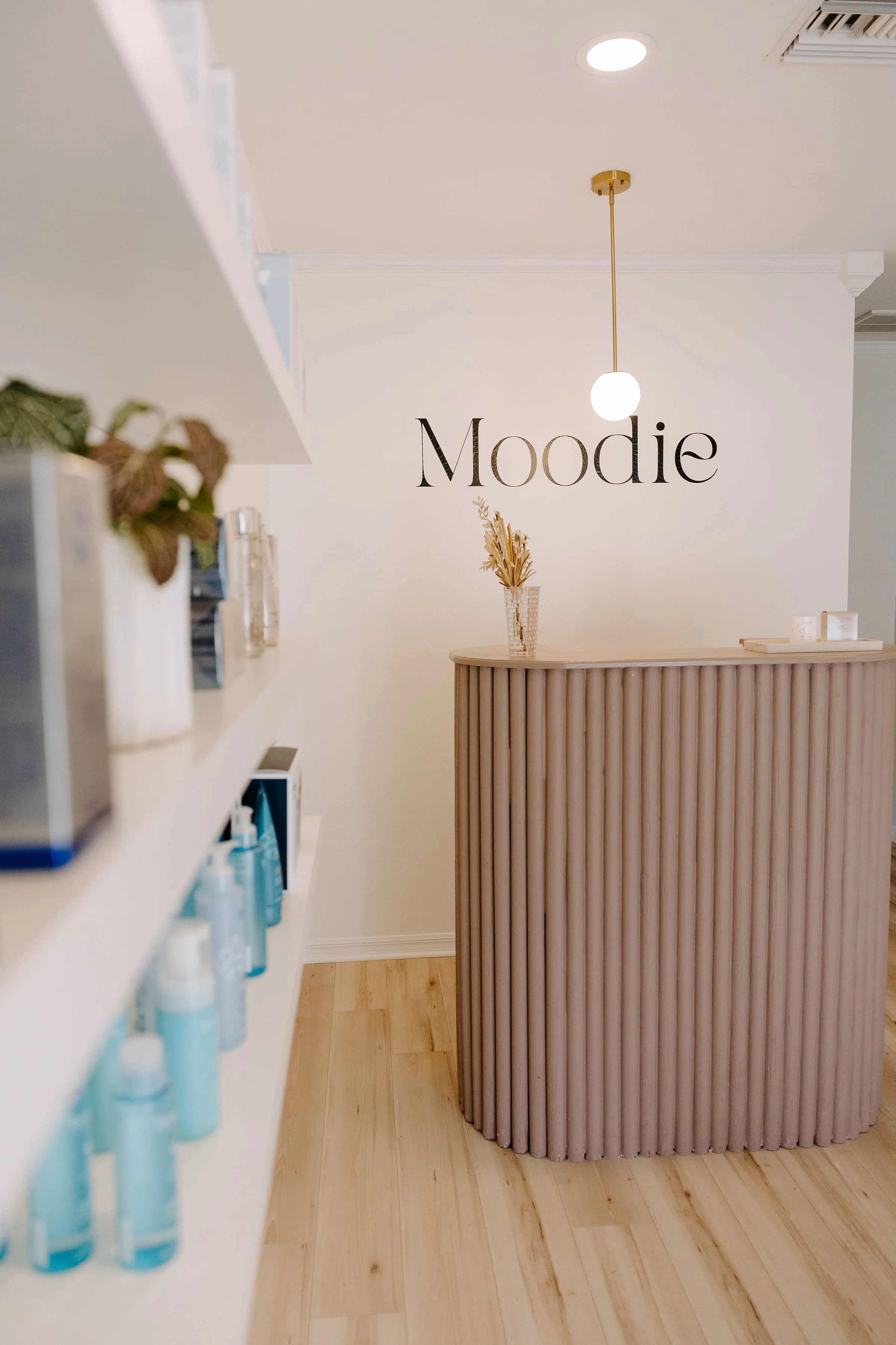 Interior of a spa or beauty salon reception area with a minimalist style, featuring a white wall with the word 'Moodie' in large black letters, a curved wooden reception desk, a small vase with dried plants on the desk, and shelves on the left with v