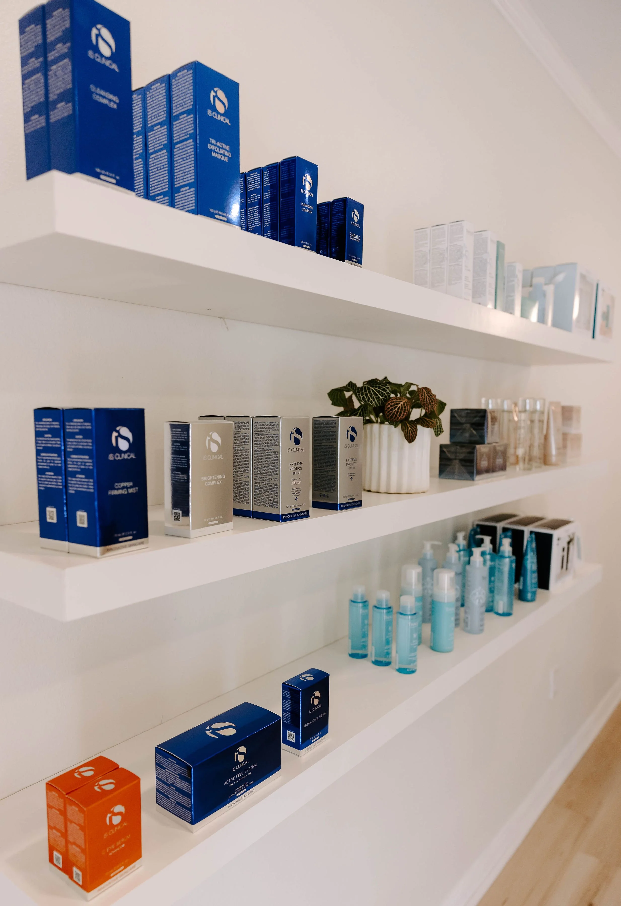 White shelves with skincare products, including blue and white boxes, a white potted plant, and blue bottles, in a skincare store or display area.