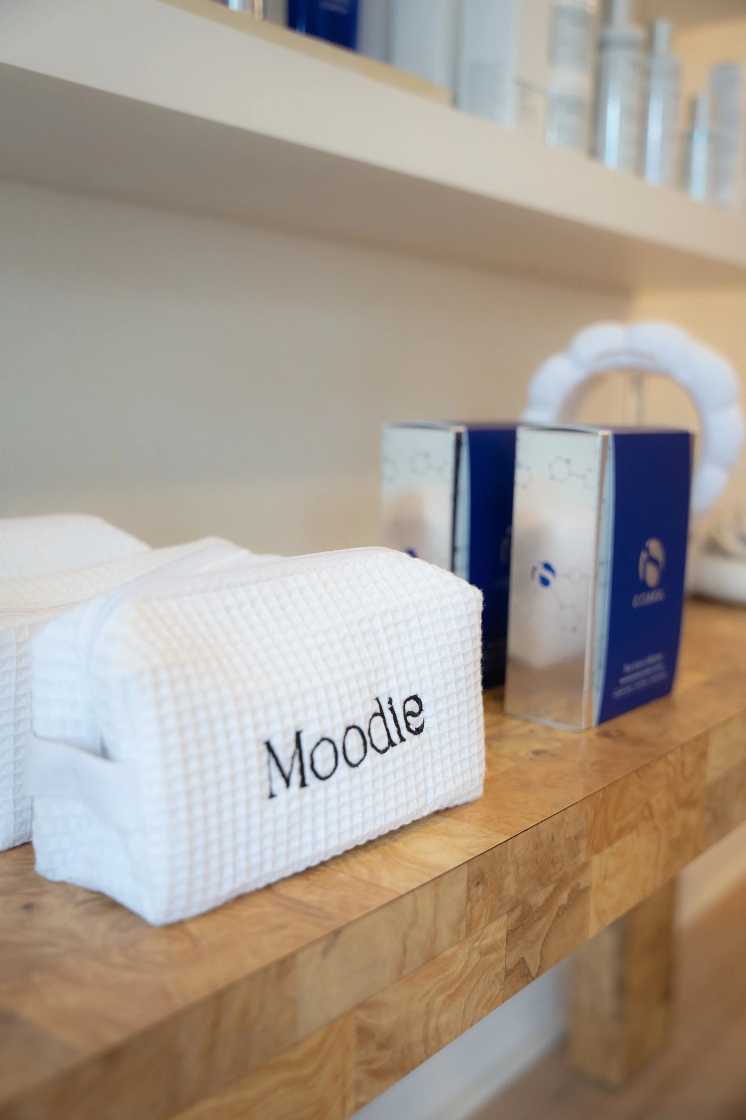 A white waffle-textured towel with the word 'Moodie' embroidered on it, placed next to a box, on a wooden surface, with a blurred background that includes a white decorative object and shelves.