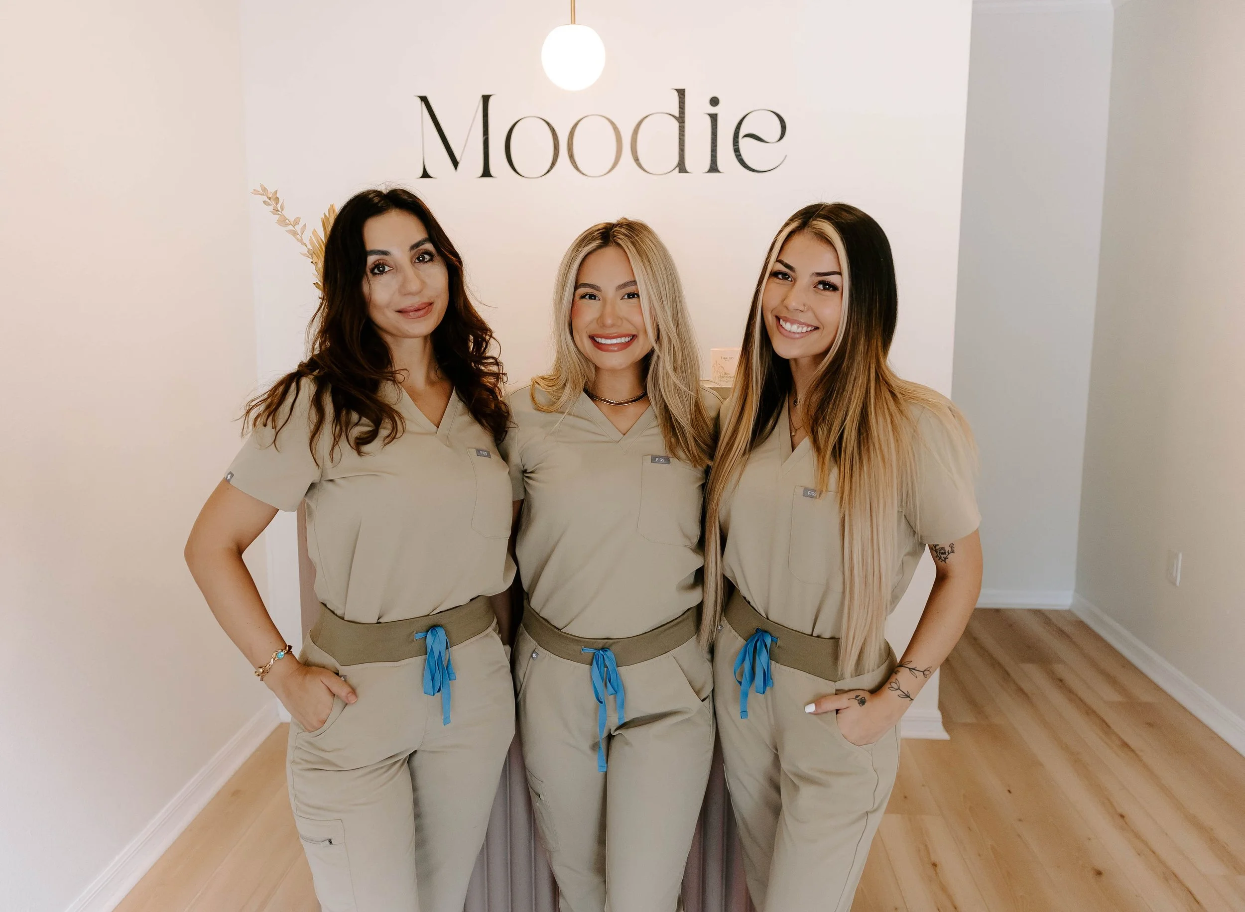 Three women in matching beige uniforms, standing closely together indoors, smiling at the camera, with a white wall behind them that reads 'Moodie'.