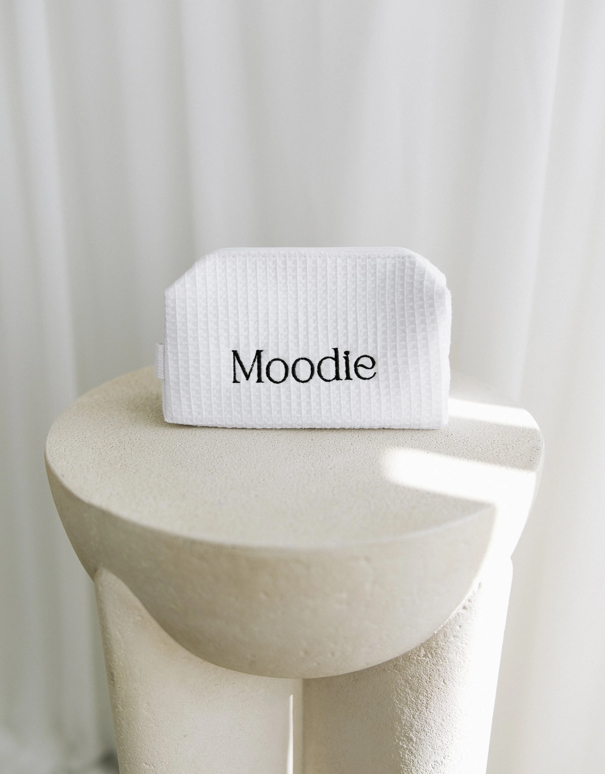 A white textured fabric pouch with the word 'Moodie' embroidered in black, placed on a light-colored, textured round surface against a plain white background.