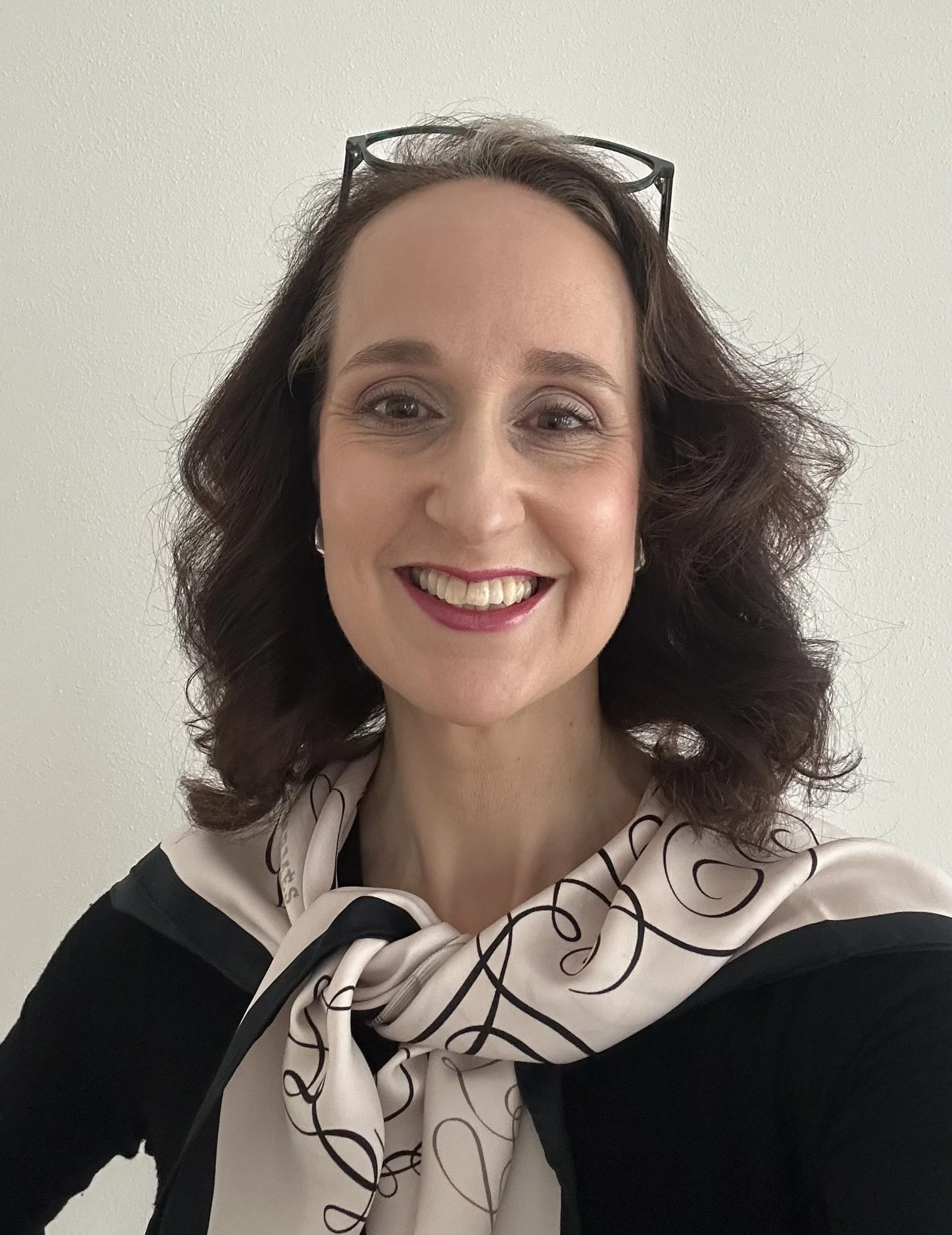 Ein fröhliches Porträt einer Frau mit braunen, lockigen Haaren, die eine Brille auf dem Kopf trägt, vor einem hellen Hintergrund.