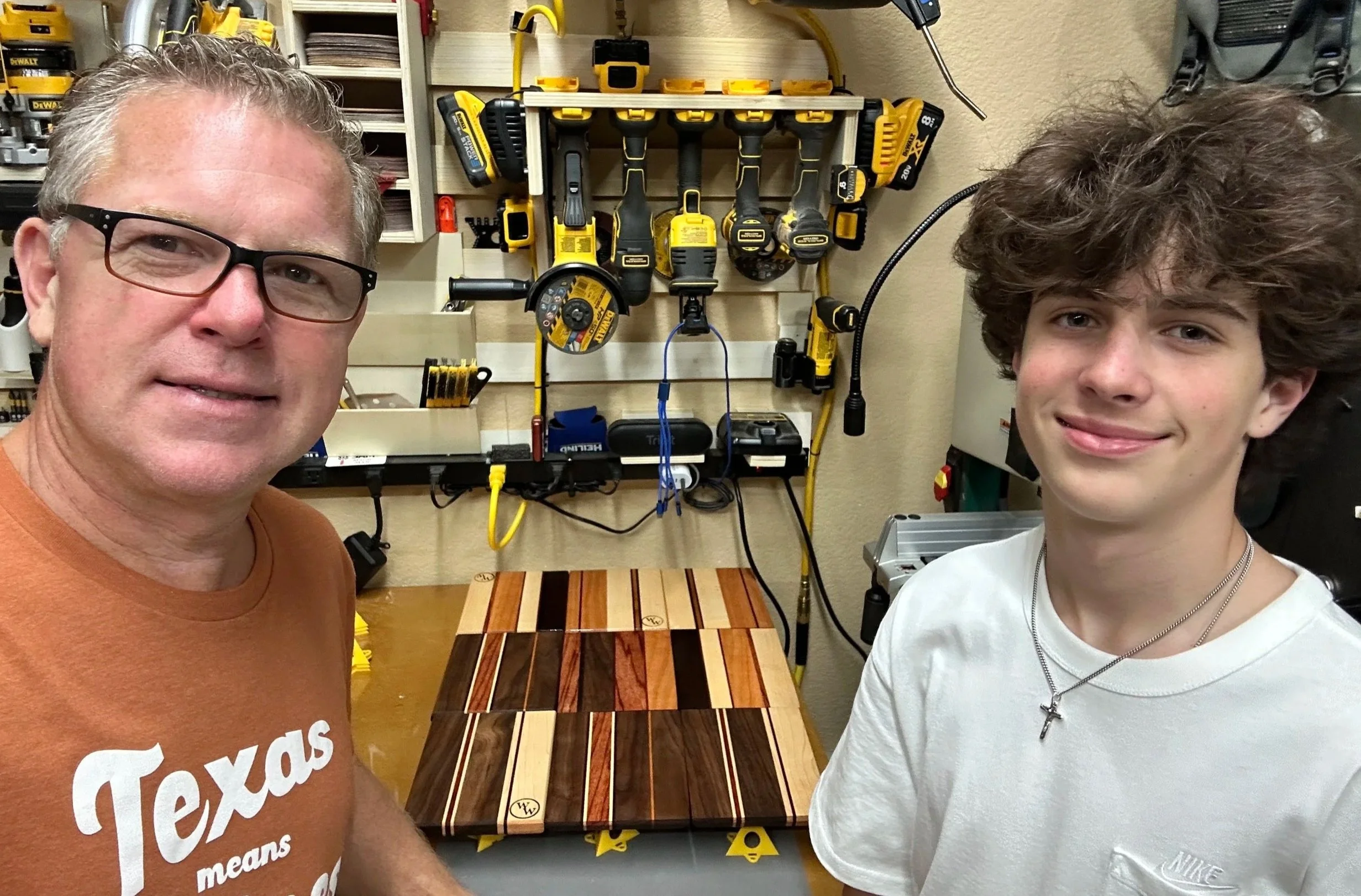 Two males standing in a workshop with a colorful wooden cutting board surface behind them and a pegboard wall with various powered tools hanging in the background.