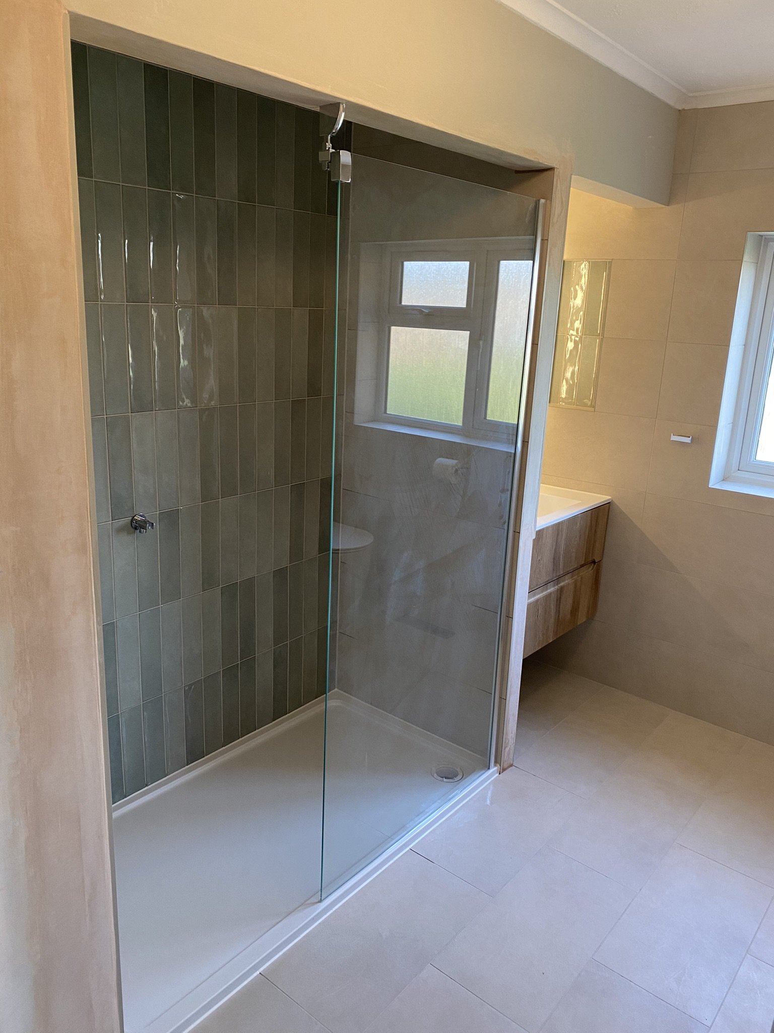 Bathroom with a walk-in shower with glass door, green tiled interior, beige floor tiles, wall-mounted wooden vanity, and a window with frosted glass.