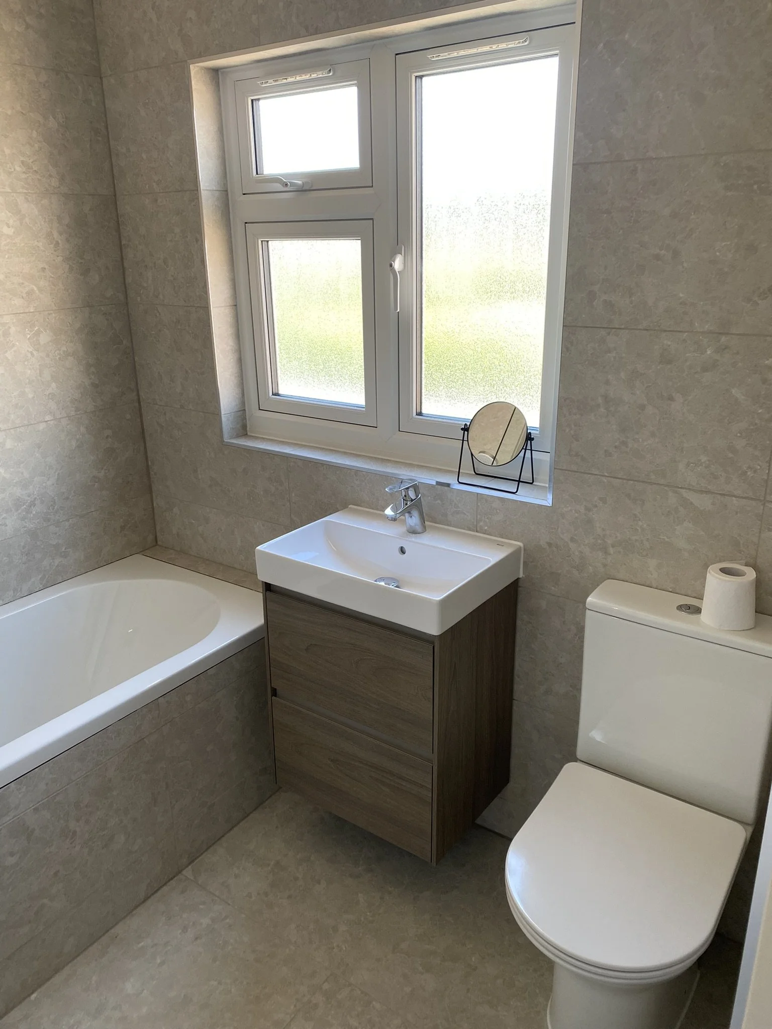 A small bathroom with frosted window, white bathtub, modern wooden vanity with sink, mirror on window sill, and toilet with a roll of toilet paper.