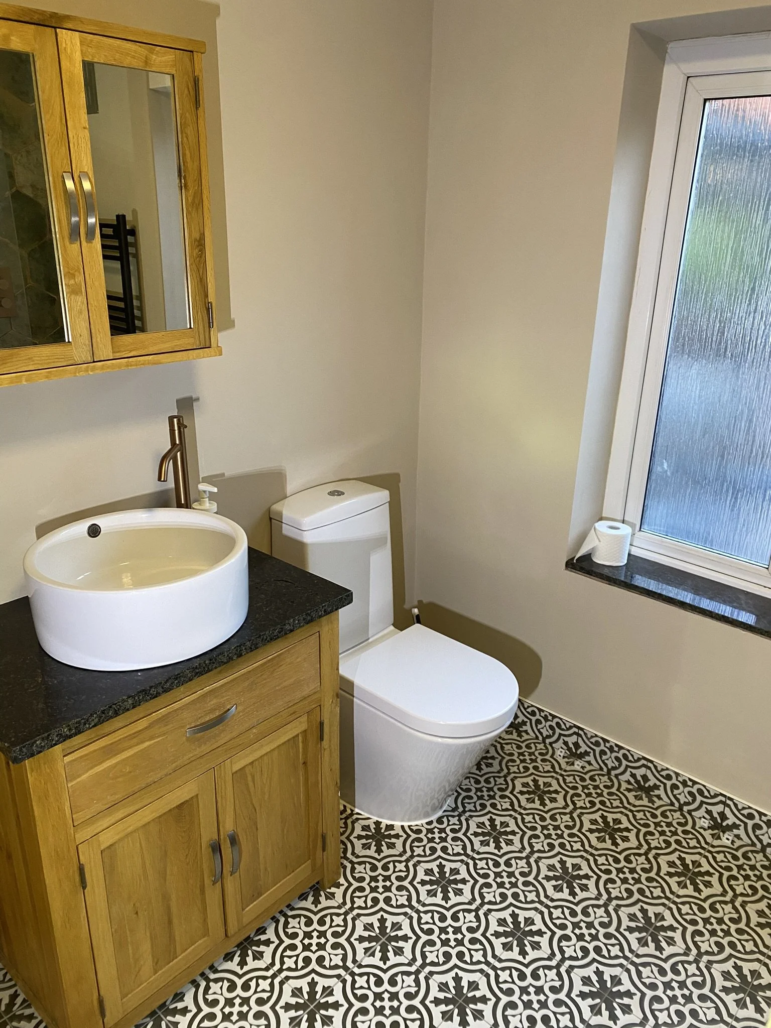 A small bathroom with a wooden cabinet with a black countertop, a white vessel sink, a modern toilet, a frosted window with a roll of toilet paper on the window sill, and patterned black and white tile flooring.