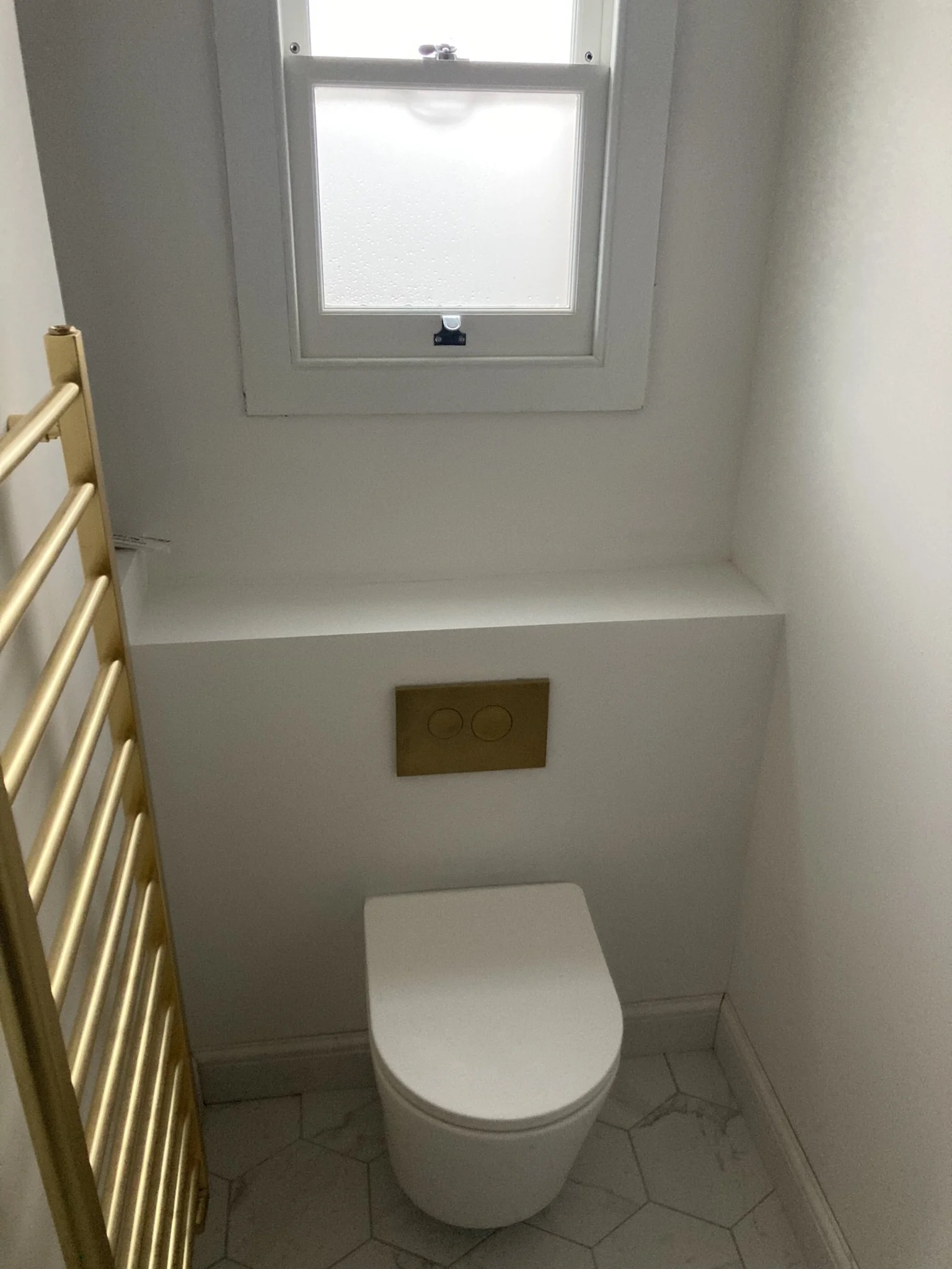 A small bathroom with a white toilet, a window above it, and a gold towel rail on the left. The floor has hexagonal tiles.