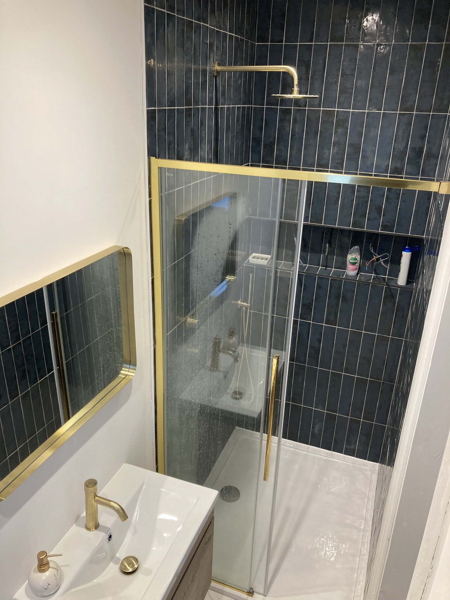 Small bathroom with a white sink, gold fixtures, glass shower with gold trim, dark blue tiled wall, and a mirror with a gold frame.