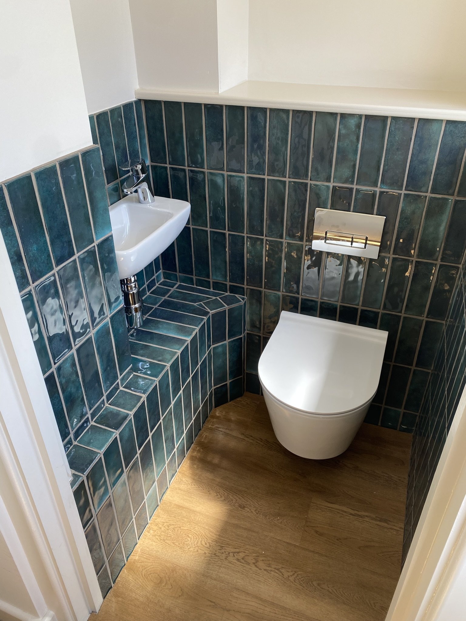 Small bathroom with teal tiled walls, a white toilet, and a tiny white sink above it, with wood-look flooring.