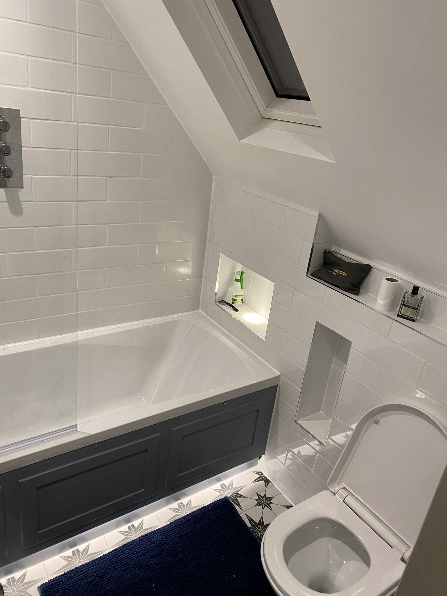 Bathroom with sloped ceiling, skylight window, toilet, bathtub with dark gray panel, decorative floor tiles, and white tiled walls. Small recessed shelf with cleaning supplies and other toiletries.