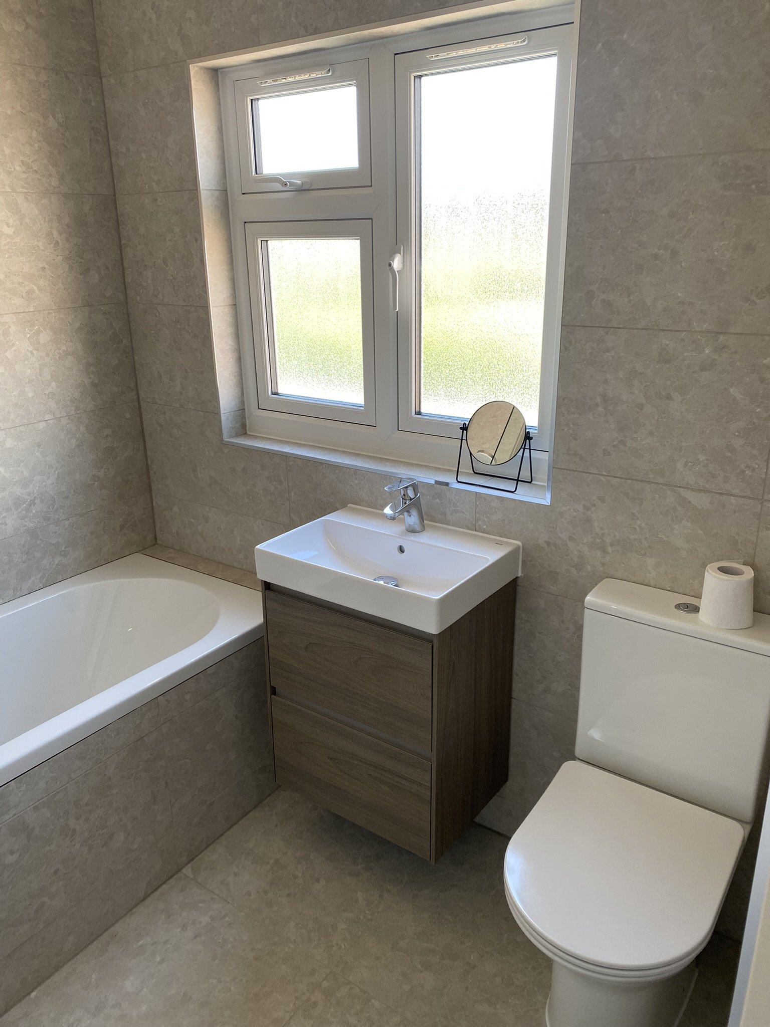 A small bathroom with a bathtub, a vanity with a sink, a mirror, and a toilet. The bathroom has beige tile walls and floor, a frosted window letting in natural light, and a roll of toilet paper on the toilet tank.