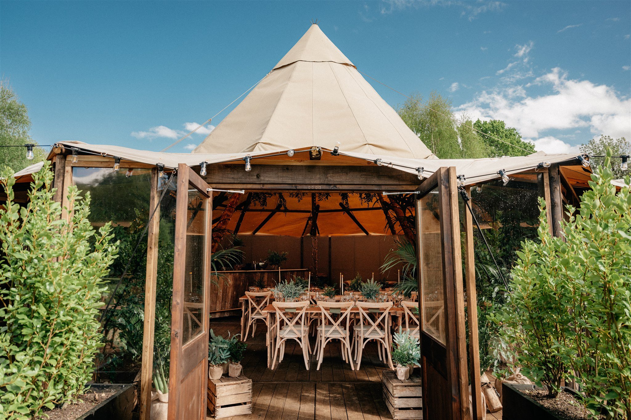 A wooden outdoor dining area under a beige tipi tent with string lights, surrounded by green bushes and trees, under a blue sky with clouds.