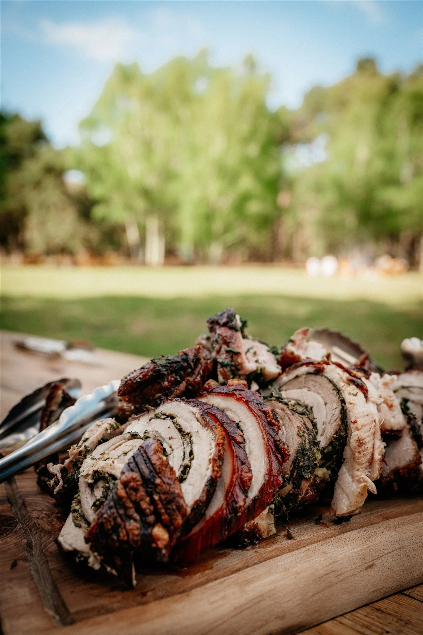 Sliced roasted meat with herbs on a wooden cutting board. Blurred trees and blue sky in the background.
