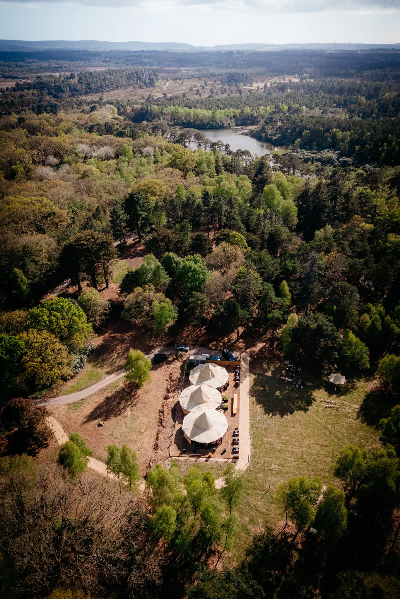 Aerial view of a park with a small lake, surrounded by dense trees with green and some bare branches, and a shaded outdoor seating area with umbrellas. In the background, there are rolling hills and forests.