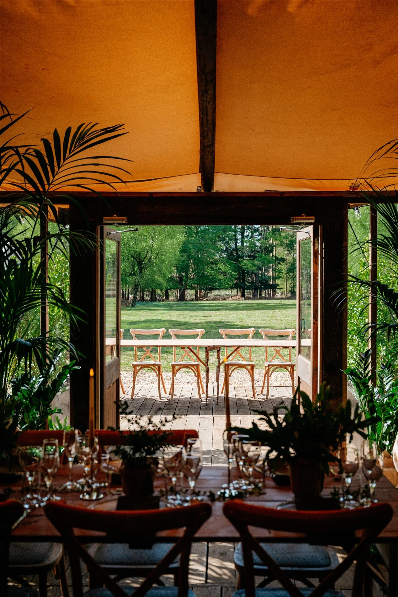 Inside view of a dining area with tables set with glasses and wine bottles, looking out through open doors to an outdoor patio with wooden chairs and a green lawn with trees.