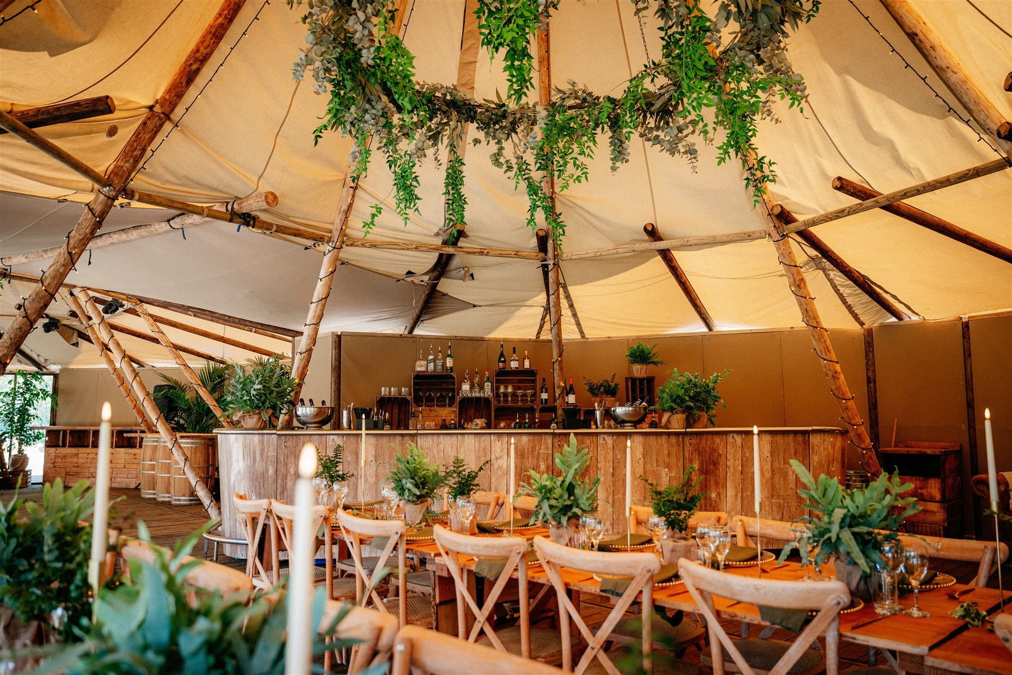 Indoor event space decorated with lush green plants and wooden furniture, featuring a bar area with bottles and glasses, under a canopy with hanging greenery.
