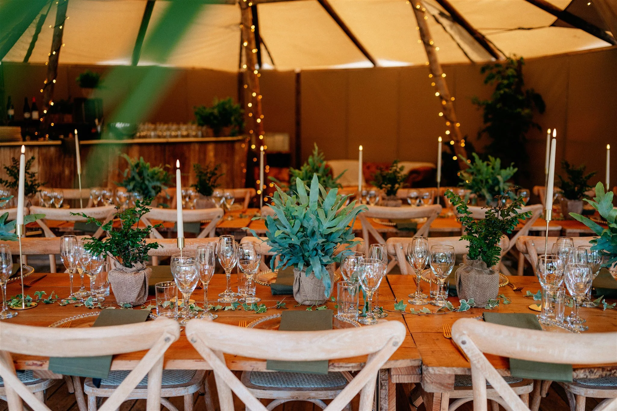 Decorated dining table with greenery in burlap-wrapped pots, surrounded by wooden chairs, set with wine glasses, candles, and napkins, in a warmly lit indoor event space with string lights and plants.