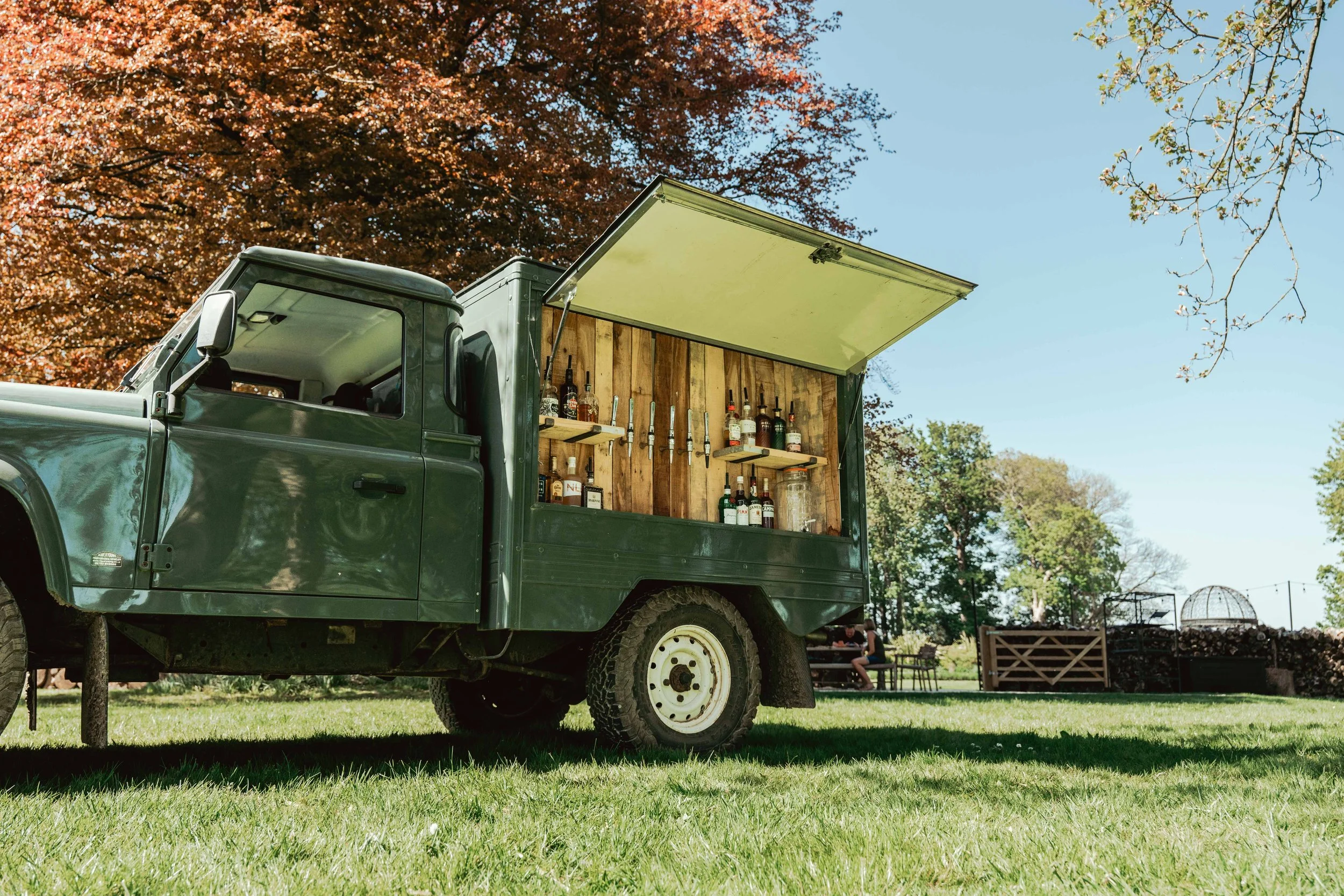 Green converted truck serving as outdoor bar with bottles and glasses inside, set on a grassy area with trees in the background.