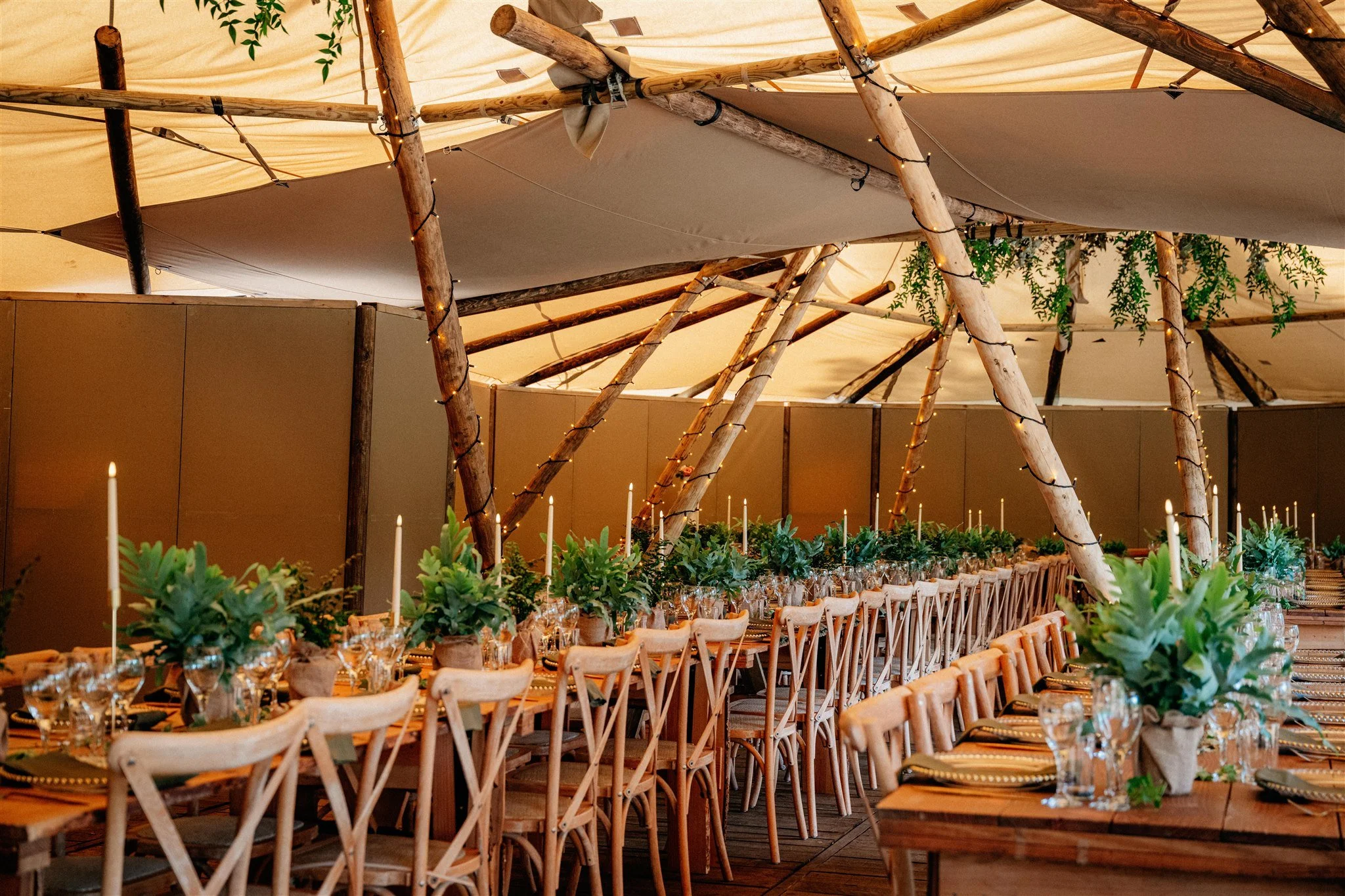Wedding reception setup with long tables, decorated with greenery and candles, under a tipi with wooden beams and draped fabric, surrounded by string lights and hanging plants.