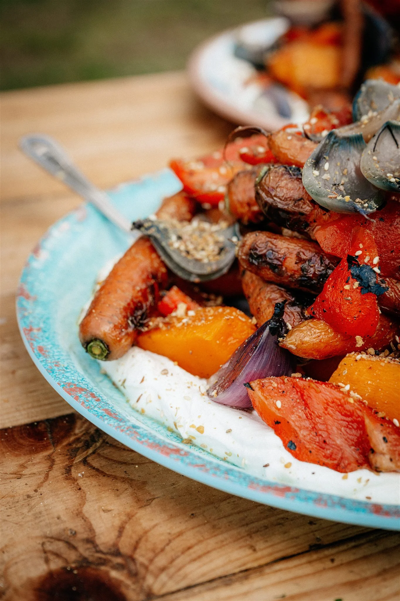 Close-up of roasted vegetables including red and yellow bell peppers, eggplant, cherry tomatoes, and garlic on a plate with a creamy dip, placed on a wooden surface.