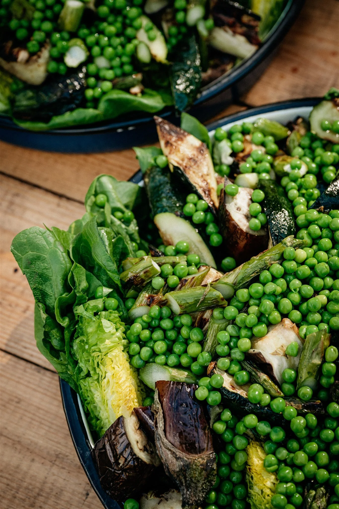 A platter of grilled vegetables including eggplant, zucchini, and leafy greens, garnished with green peas on a wooden table.