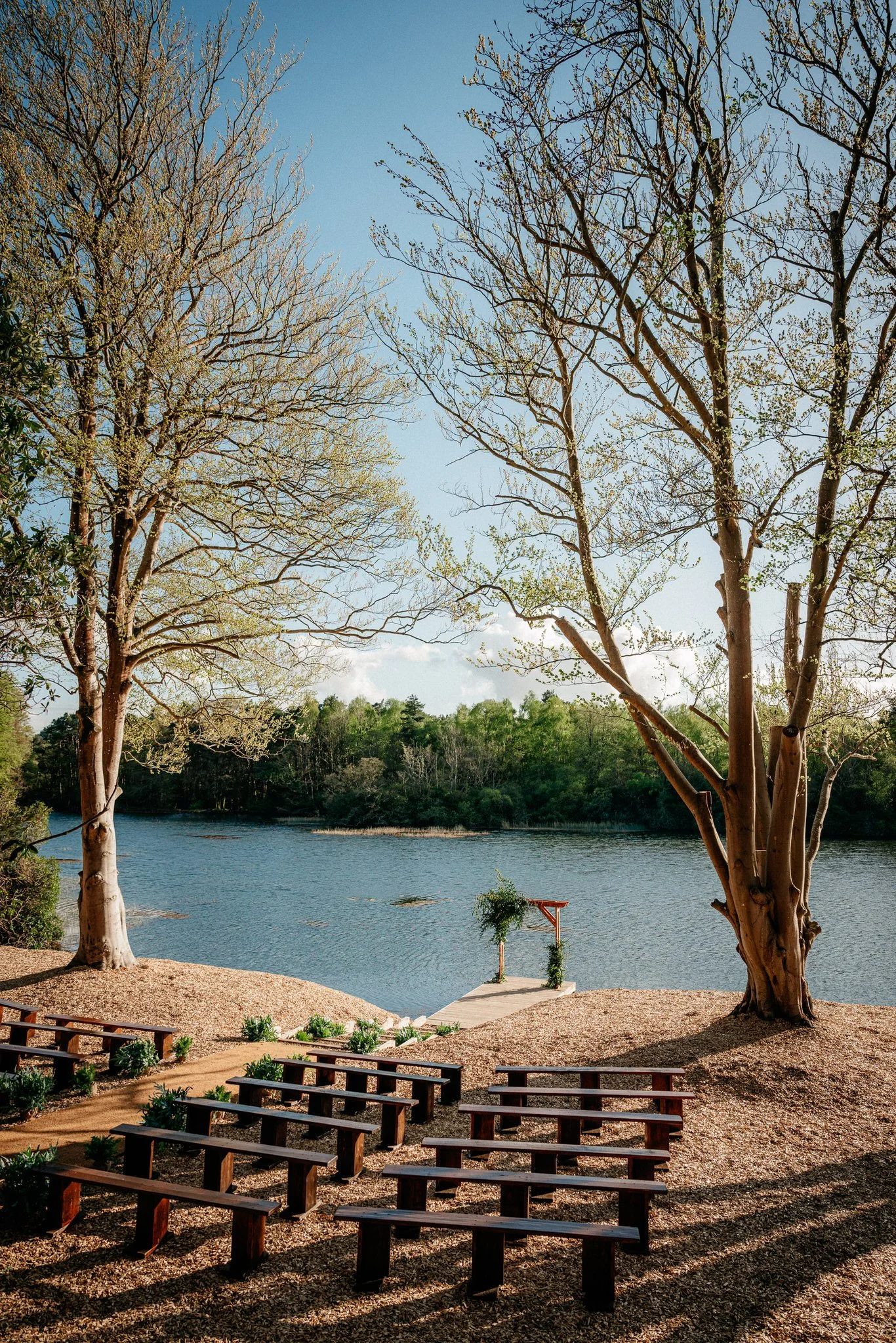 Outdoor wedding altar by a lake with two large trees and a clear blue sky.
