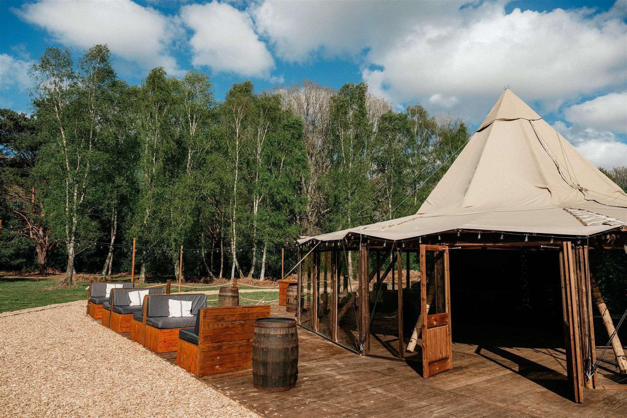 Outdoor seating area with wooden couches and barrels, next to a large canvas tent with string lights, set against a backdrop of tall green trees and a partly cloudy sky.