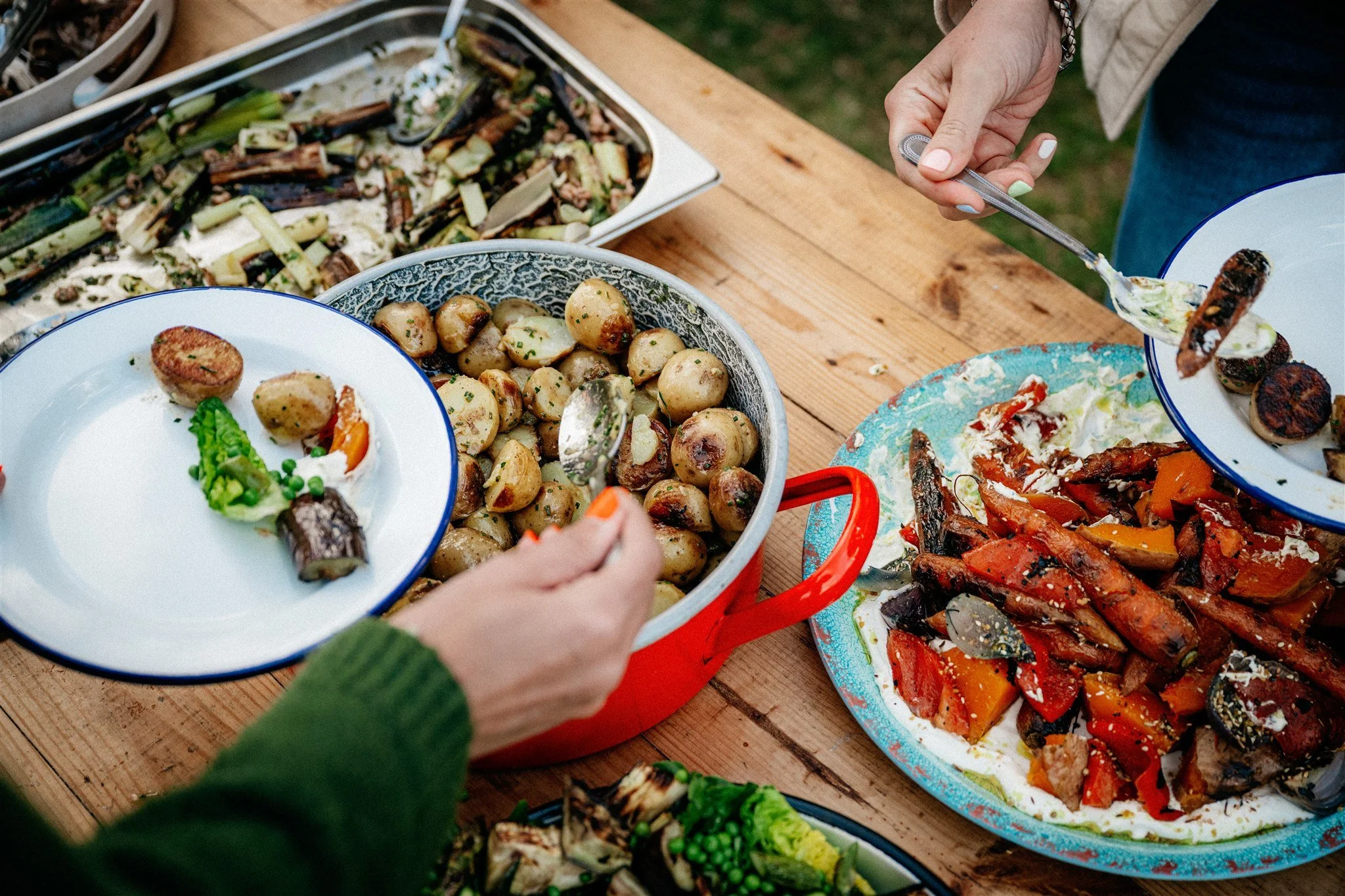 Person serving grilled vegetables, including zucchini and eggplant, at an outdoor meal with various dishes on a wooden table.
