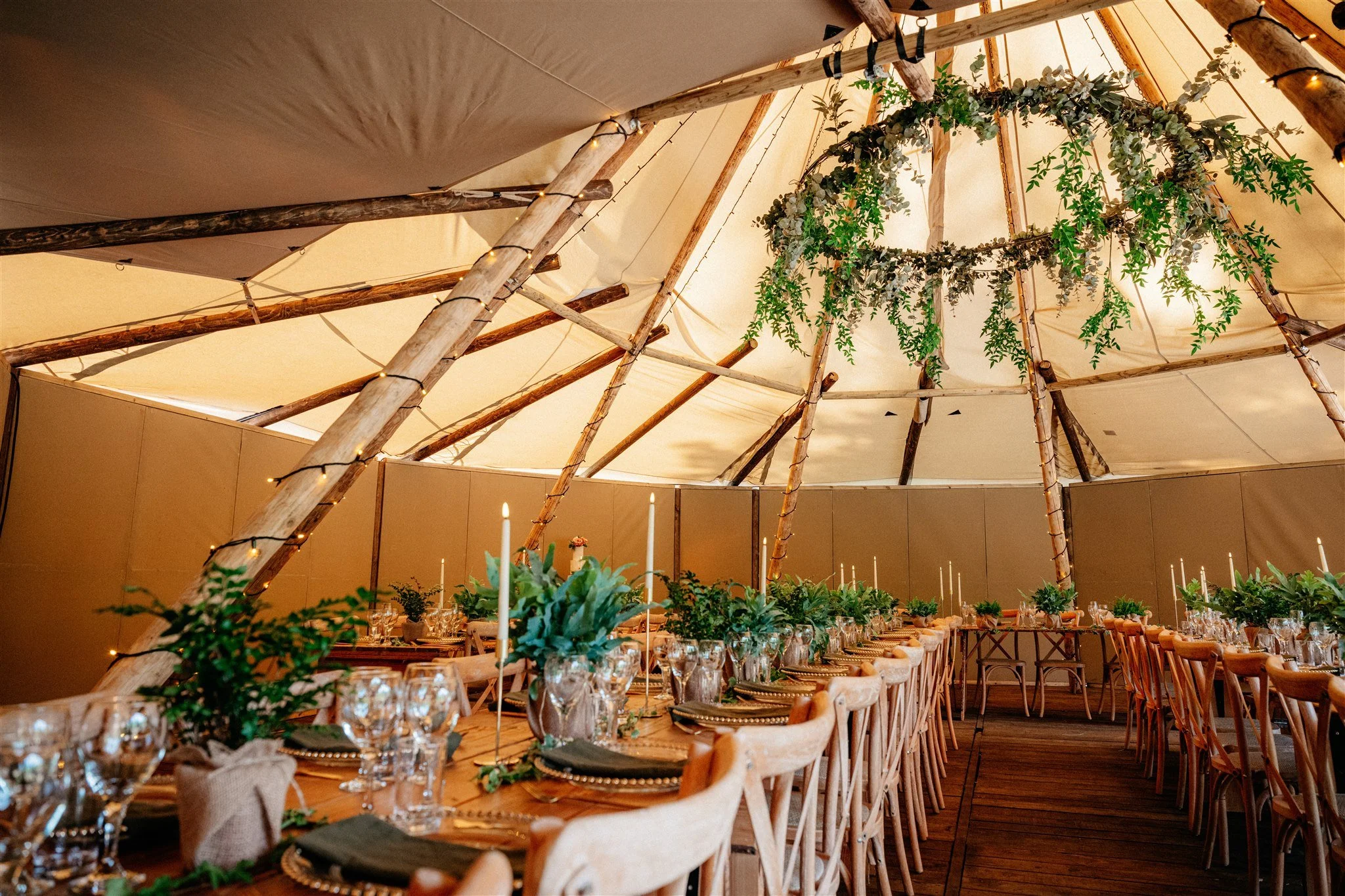 Indoor wedding reception setup with long banquet tables decorated with green foliage and candles. The ceiling is decorated with draped fabric, string lights, and hanging greenery, creating a rustic, elegant ambiance.