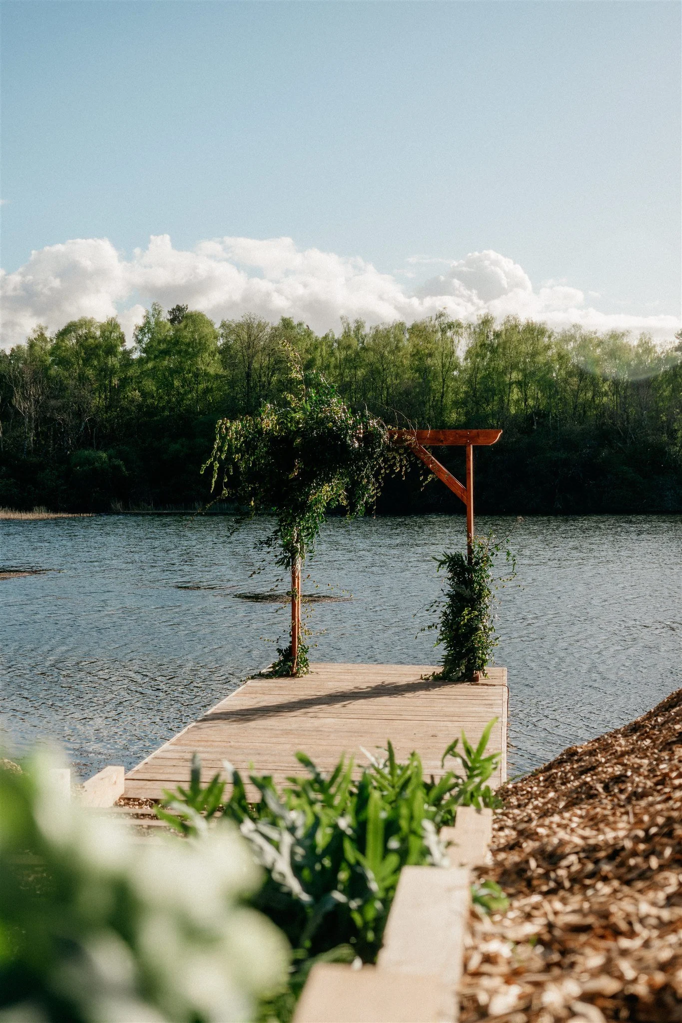Wooden dock extending over a body of water with a greenery archway at the end, surrounded by trees and cloudy sky.