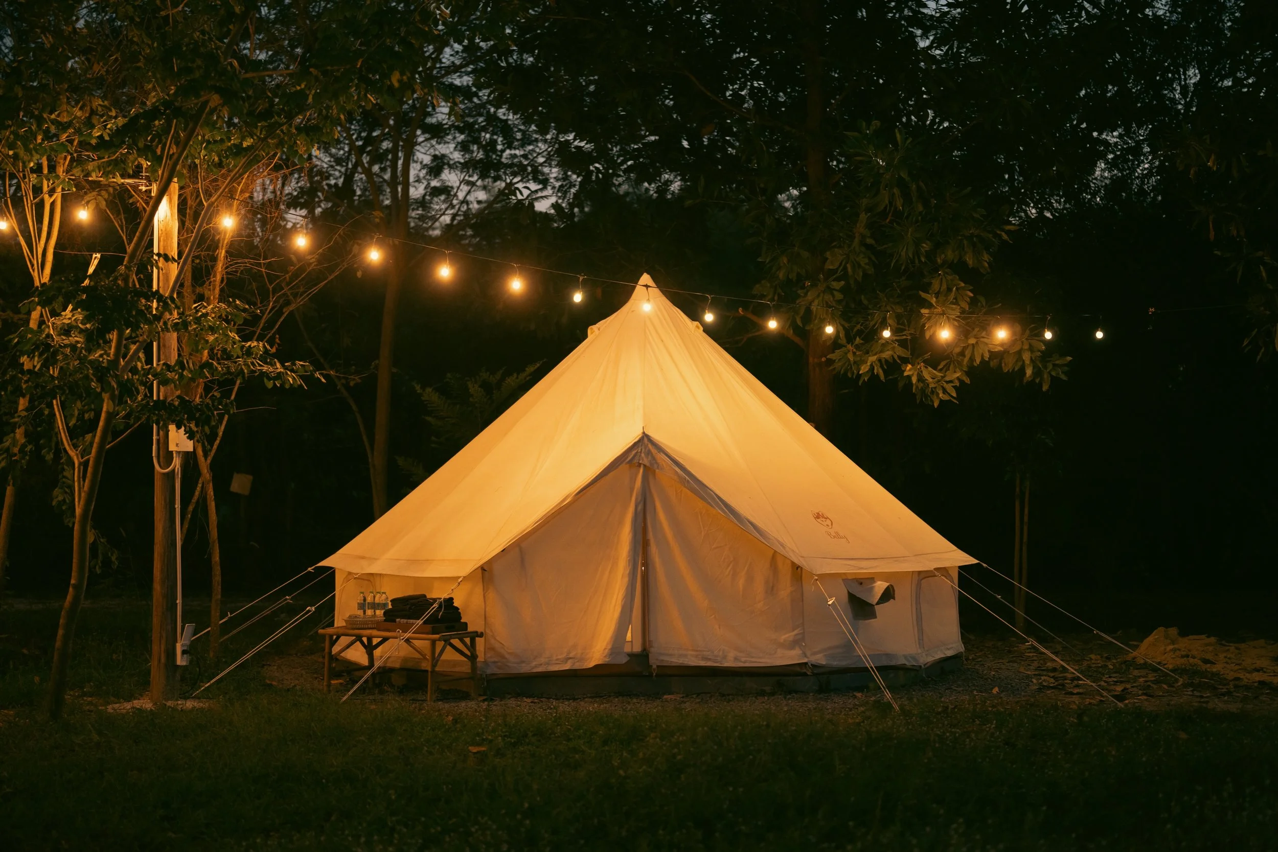 A large beige camping tent illuminated by string lights hanging above it, set in a wooded outdoor area at night.