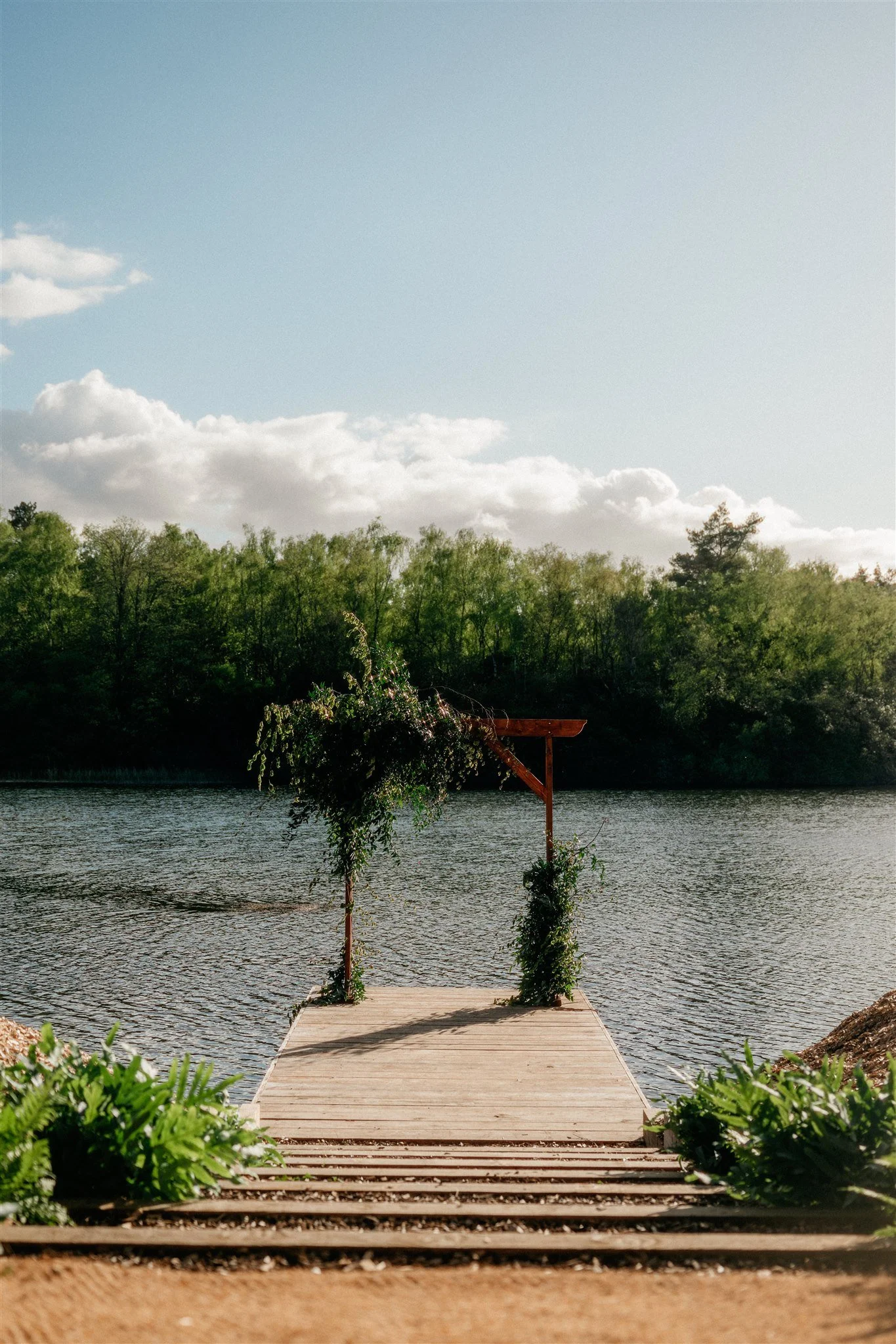 A wooden dock extending over a body of water with a decorated archway at the end, surrounded by greenery, with trees and a partly cloudy sky in the background.