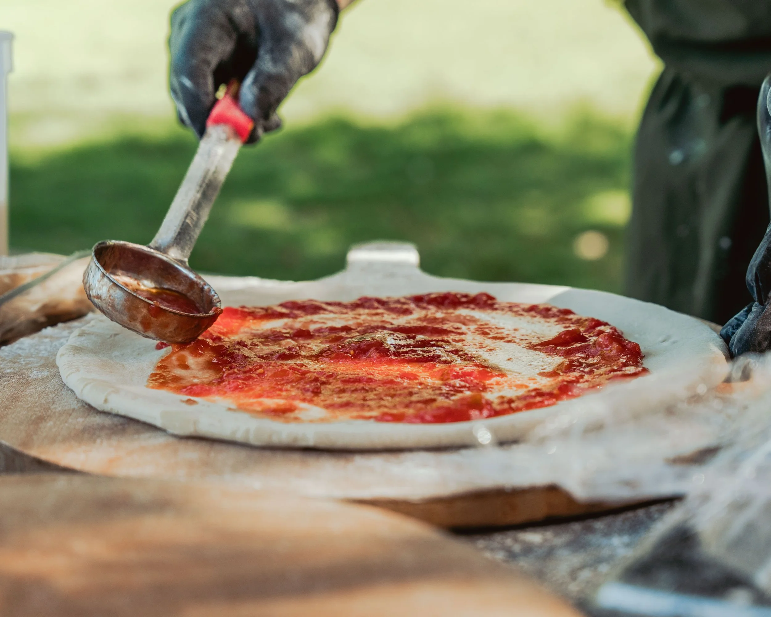 Person spreading tomato sauce on pizza dough with a ladle outdoors.