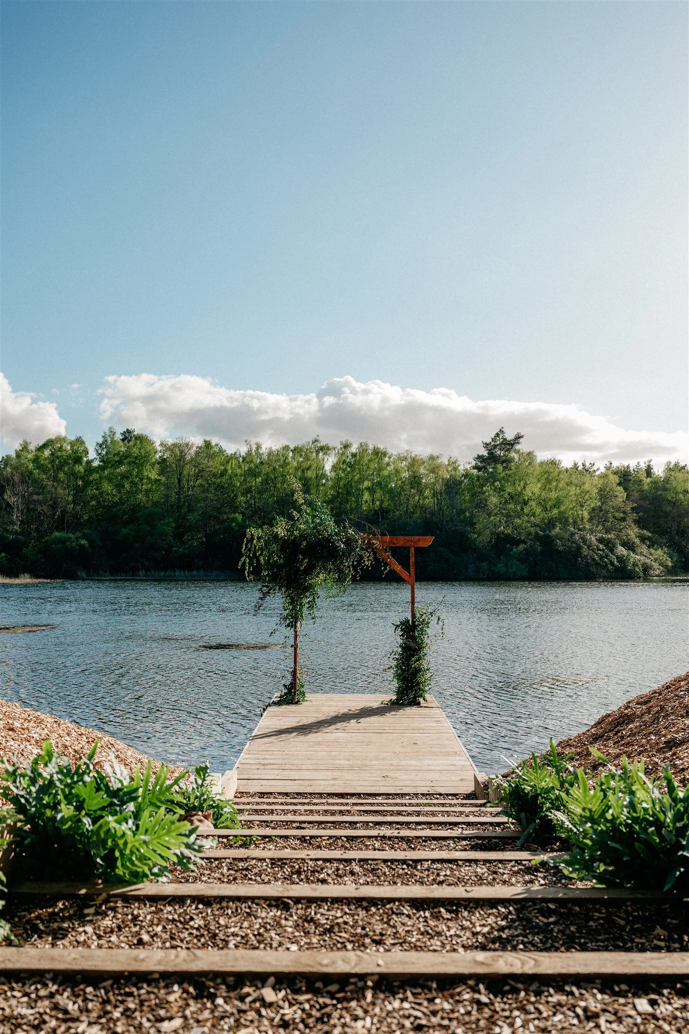 Wooden dock extending over a body of water, with two trees and greenery at the entrance, leading to a small pier with a decorative archway. The scene is set in a natural environment with cloudy sky and lush green trees in the background.