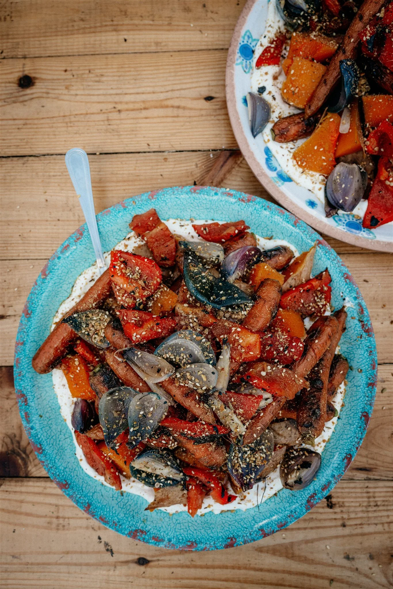 A close-up of two pizzas topped with roasted vegetables including red peppers, butternut squash, eggplant, red onion, and sprinkled with sesame seeds, served on a wooden table.