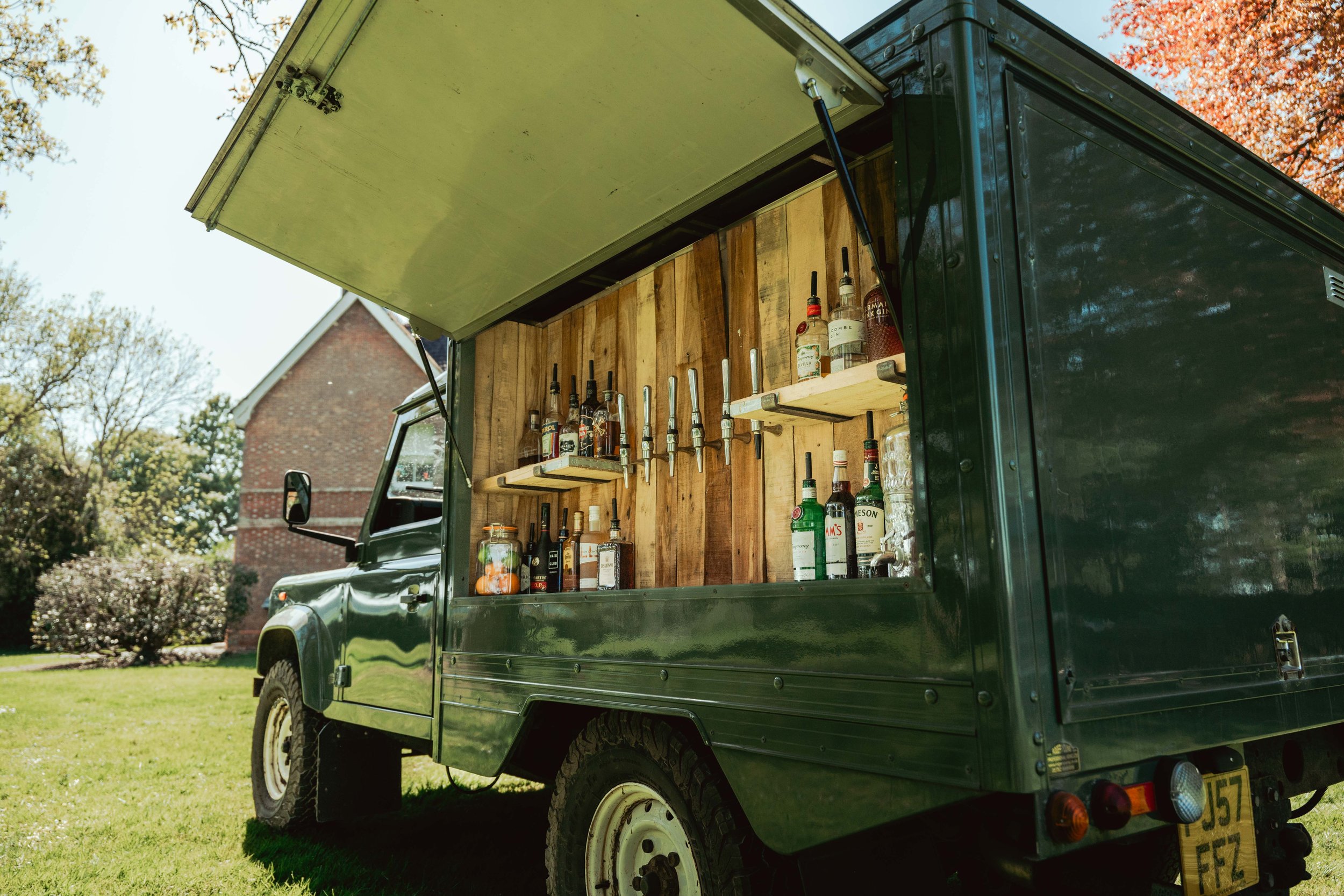 A mobile bar setup inside a dark green truck with a wooden interior, featuring shelves of liquor bottles and taps for serving drinks, parked on a grassy area with trees and a building in the background.