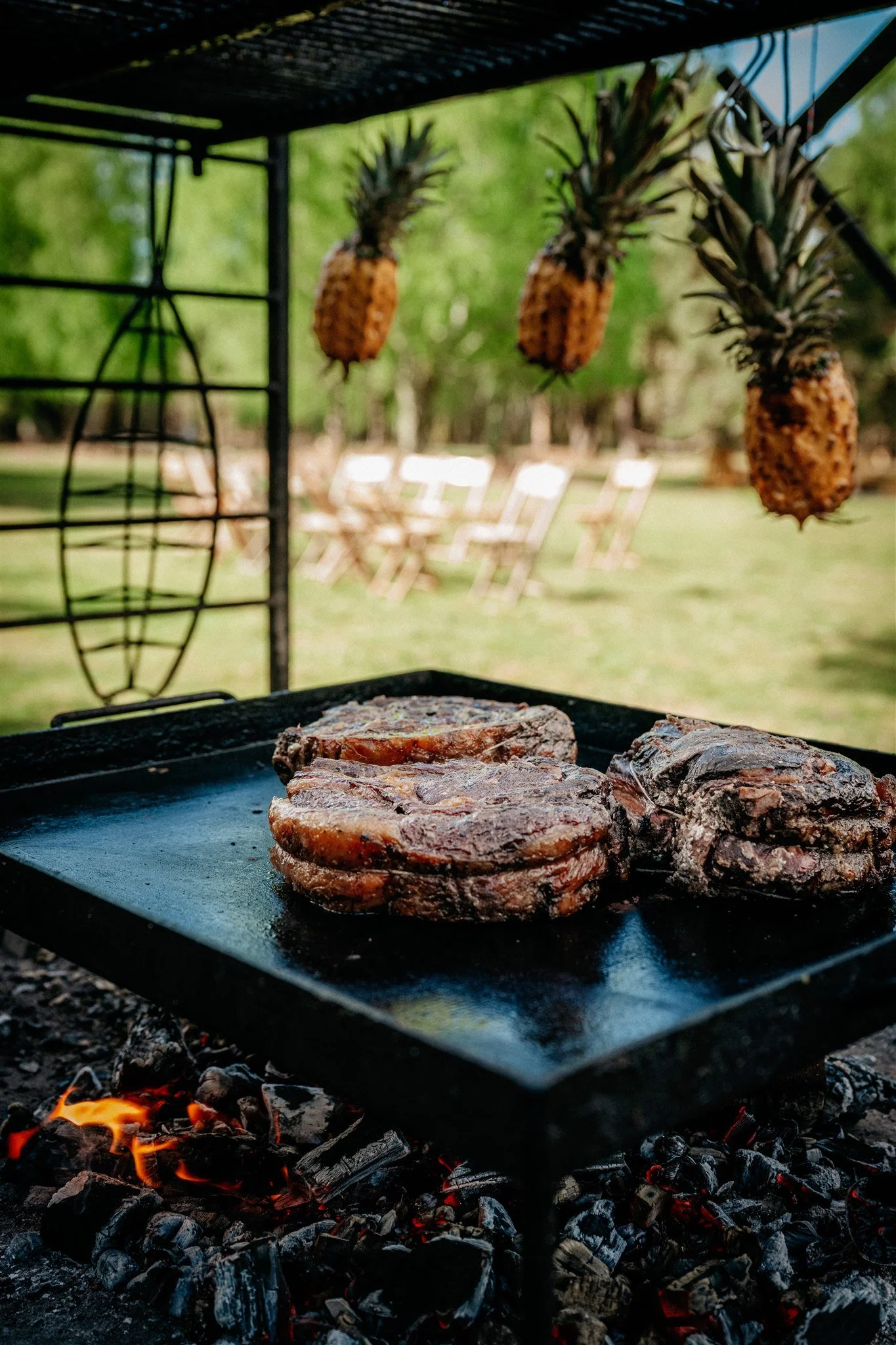 Three pieces of seasoned meat cooking on a black flat grill over hot charcoal with orange flames, pineapples hanging above in the background, and outdoor chairs blurred in the distance.